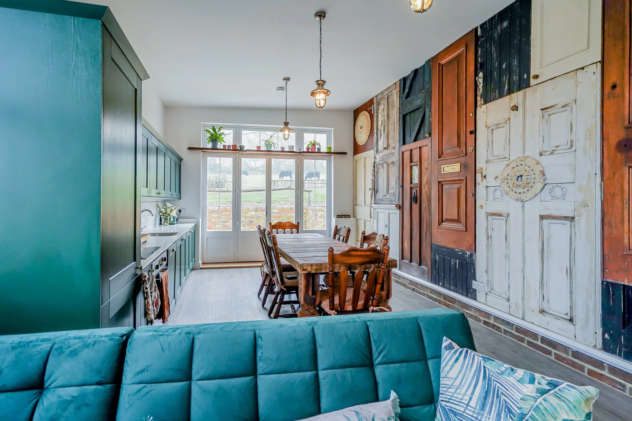 Interior of a dining room with a rustic wall of various wood panels, a wooden dining table, and chairs. A blue-green couch with pillows partially visible in the foreground. Large window and glass door leading outside with plants and a view of a field