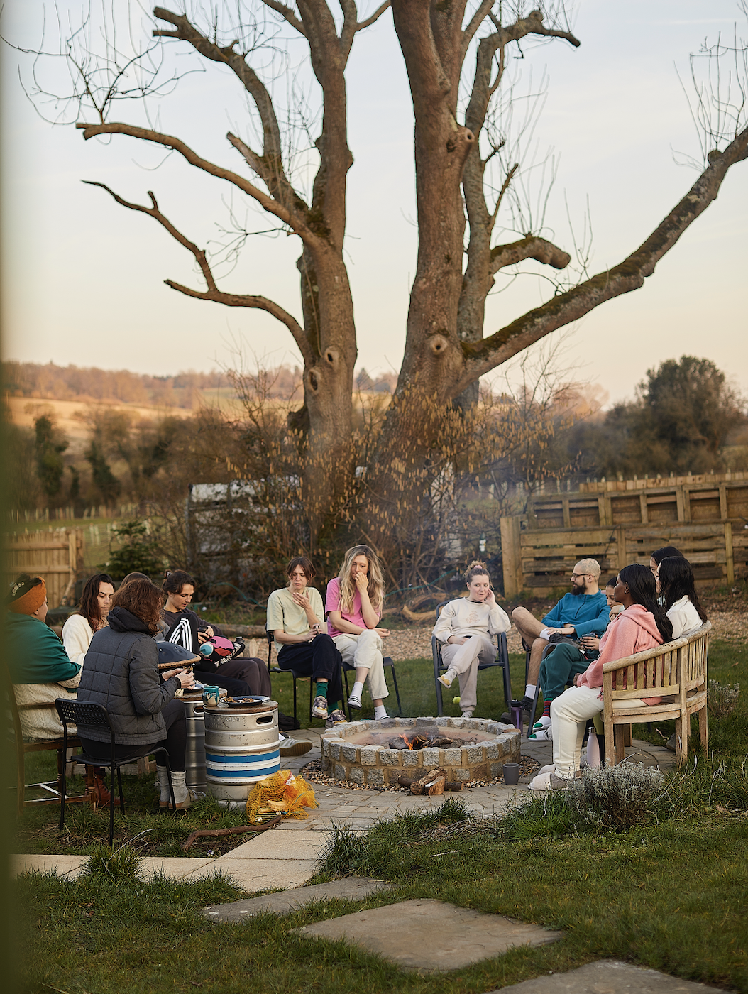 A group of people on a Yoga retreat gathered around a fire pit in a backyard during late afternoon or early evening, seated on various chairs and benches, with a large leafless tree and a wooden fence in the background.