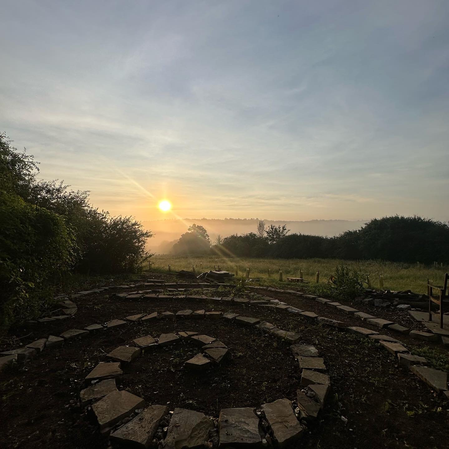 Sunrise over a misty field with a stone spiral path and surrounding greenery.