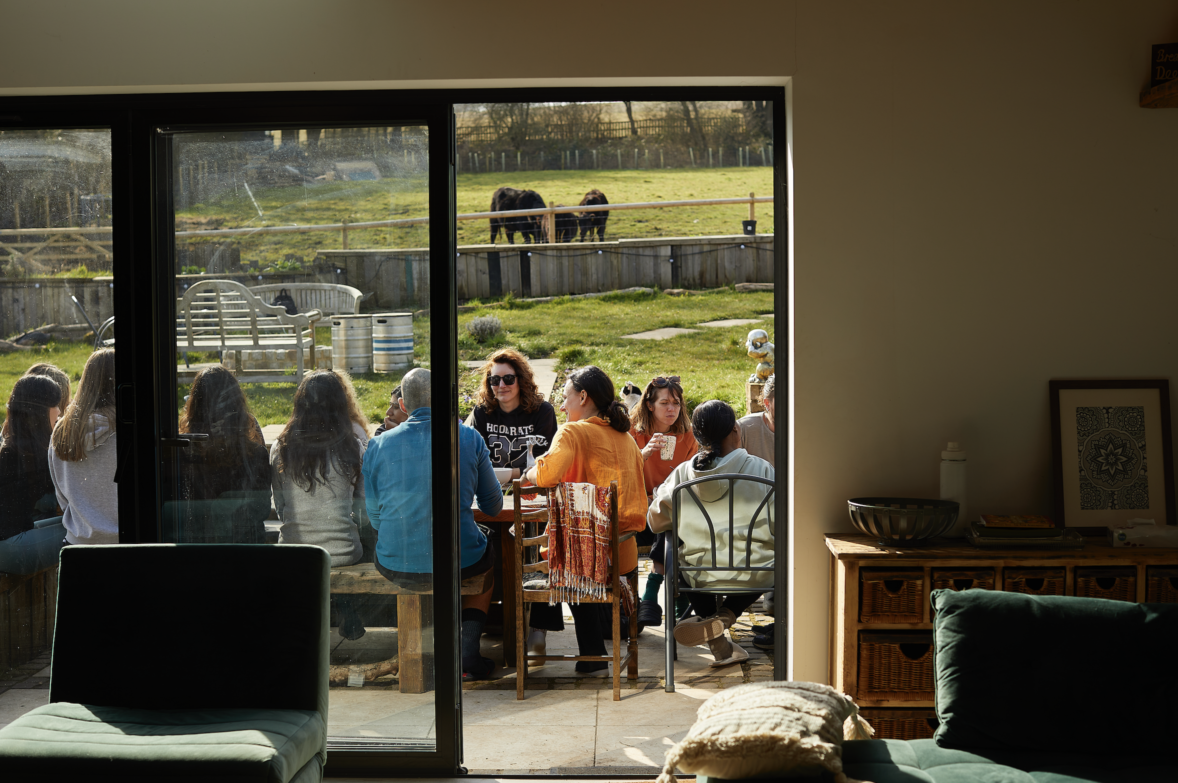 People sitting at a table outside, viewed through a sliding glass door, with a grassy field and horses in the background.