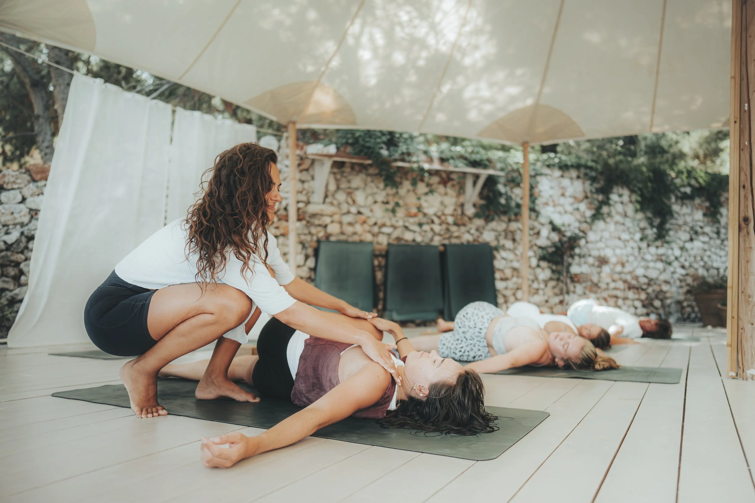 A woman in a brown top and black shorts is receiving a seated yoga pose assistance from an instructor in a white top and black shorts, with other people practicing yoga on mats under a canopy outdoors.