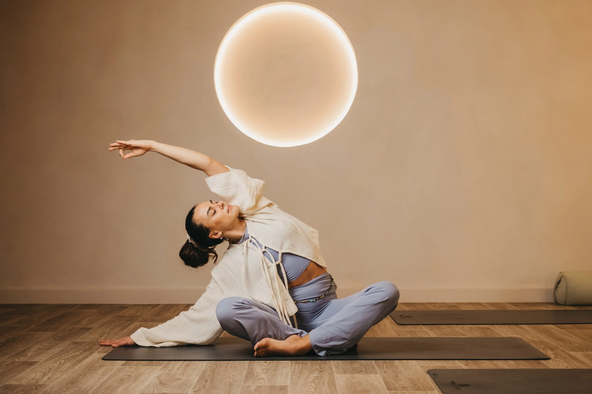 A woman practicing yoga or stretching on a mat in a room with a beige wall and a circular light fixture.