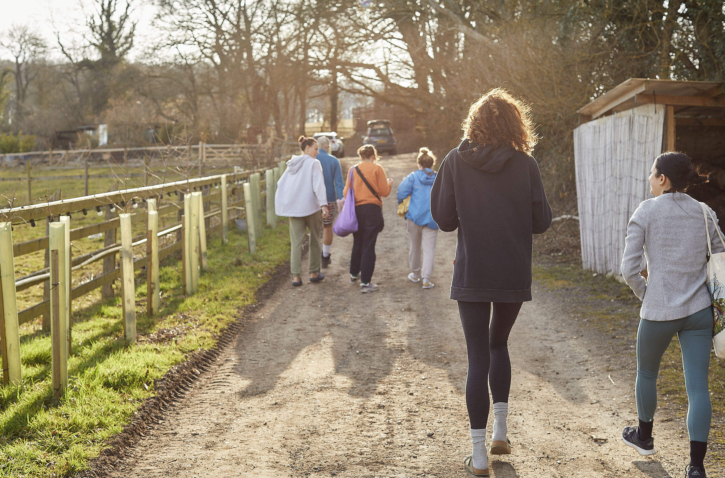 Group of people walking on a dirt path in a rural area with fences and trees, some carrying bags, during sunset or late afternoon.