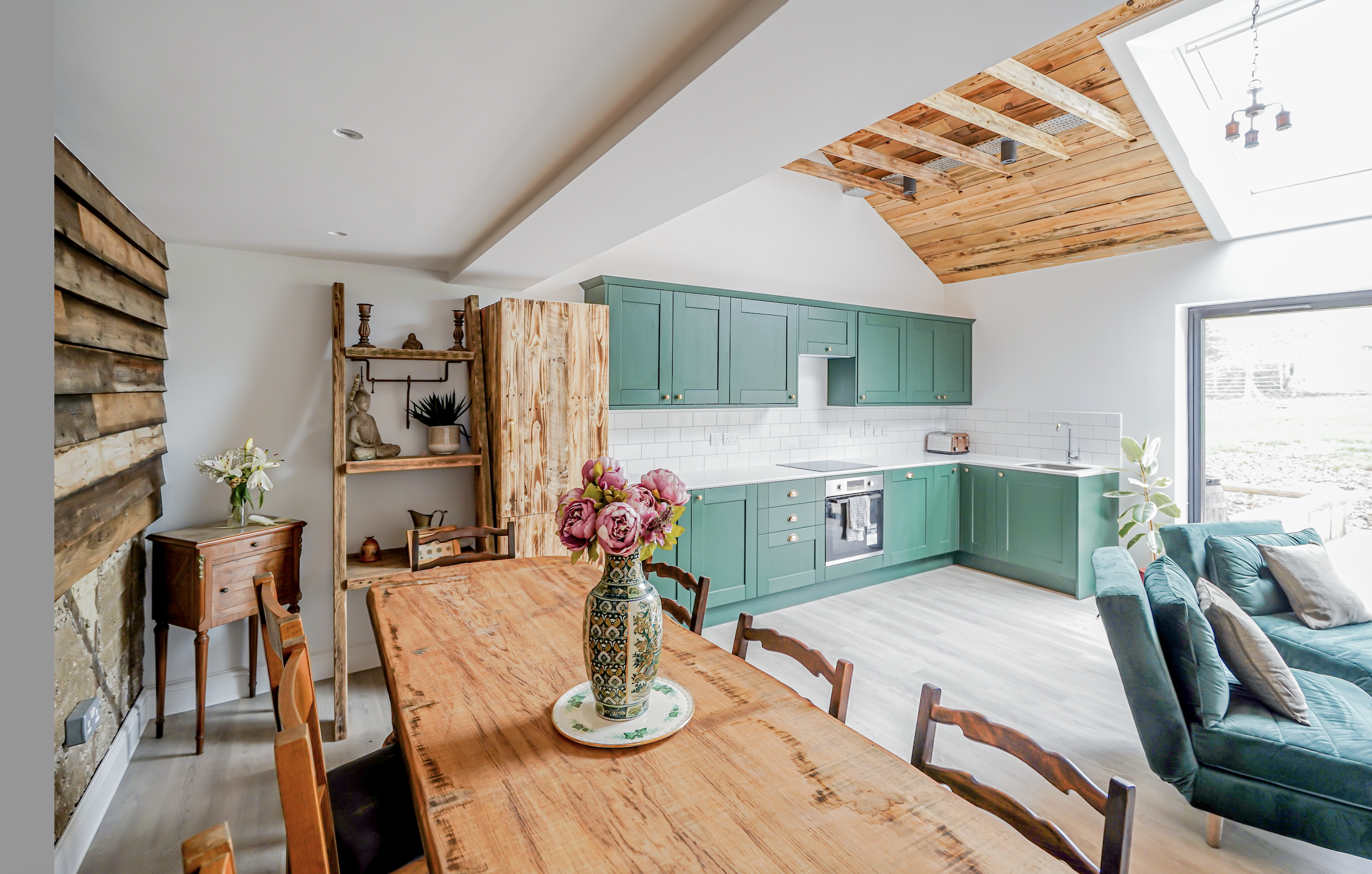 Open-plan kitchen and dining area with green cabinetry, a wooden dining table with a flower vase, and a sitting area with a teal sofa. The space features a wooden vaulted ceiling with a skylight.