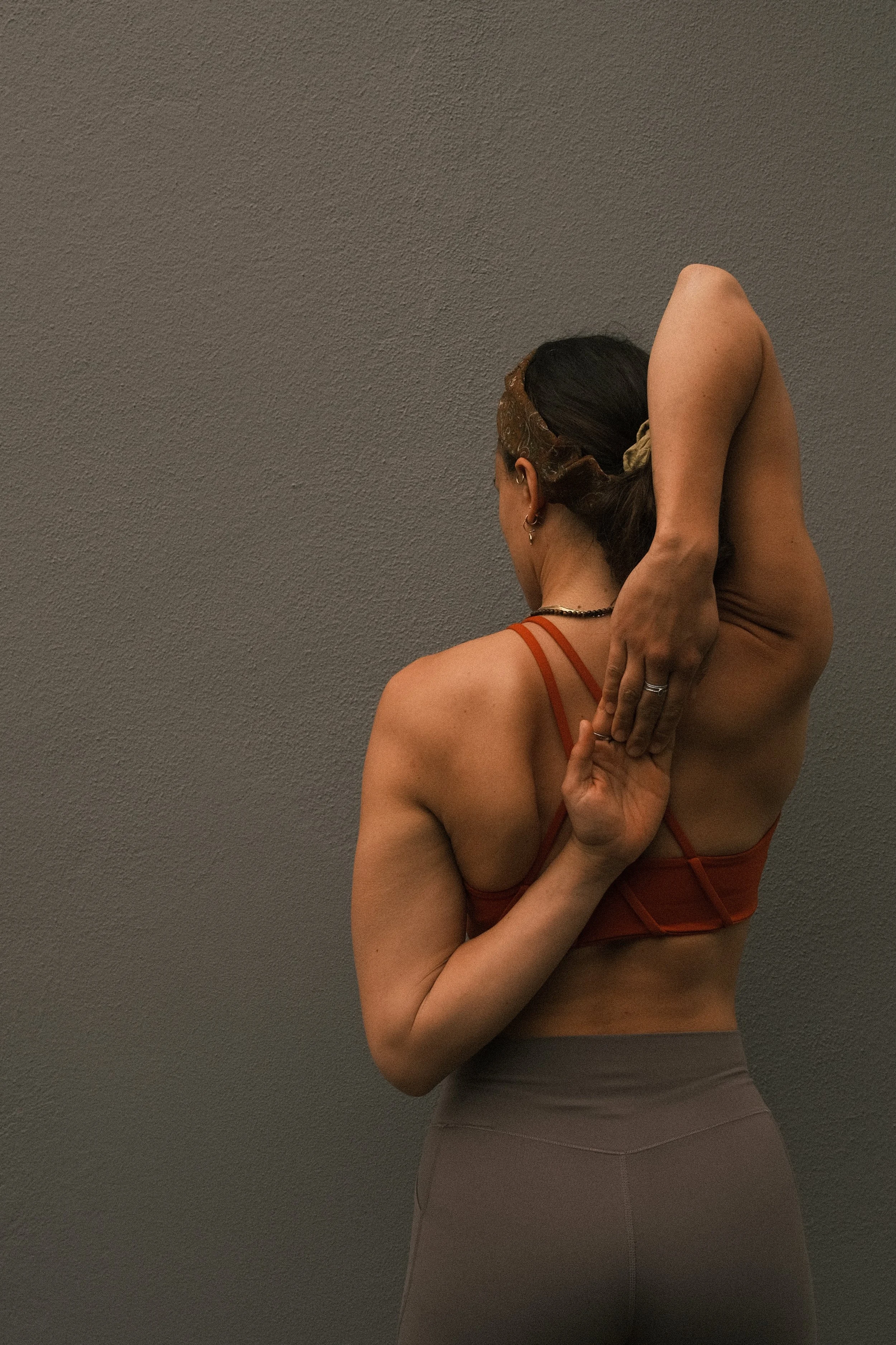 A woman practicing yoga, standing against a plain wall, in a pose with one arm bent behind her head and the other arm reaching behind her back, wearing athletic clothing.
