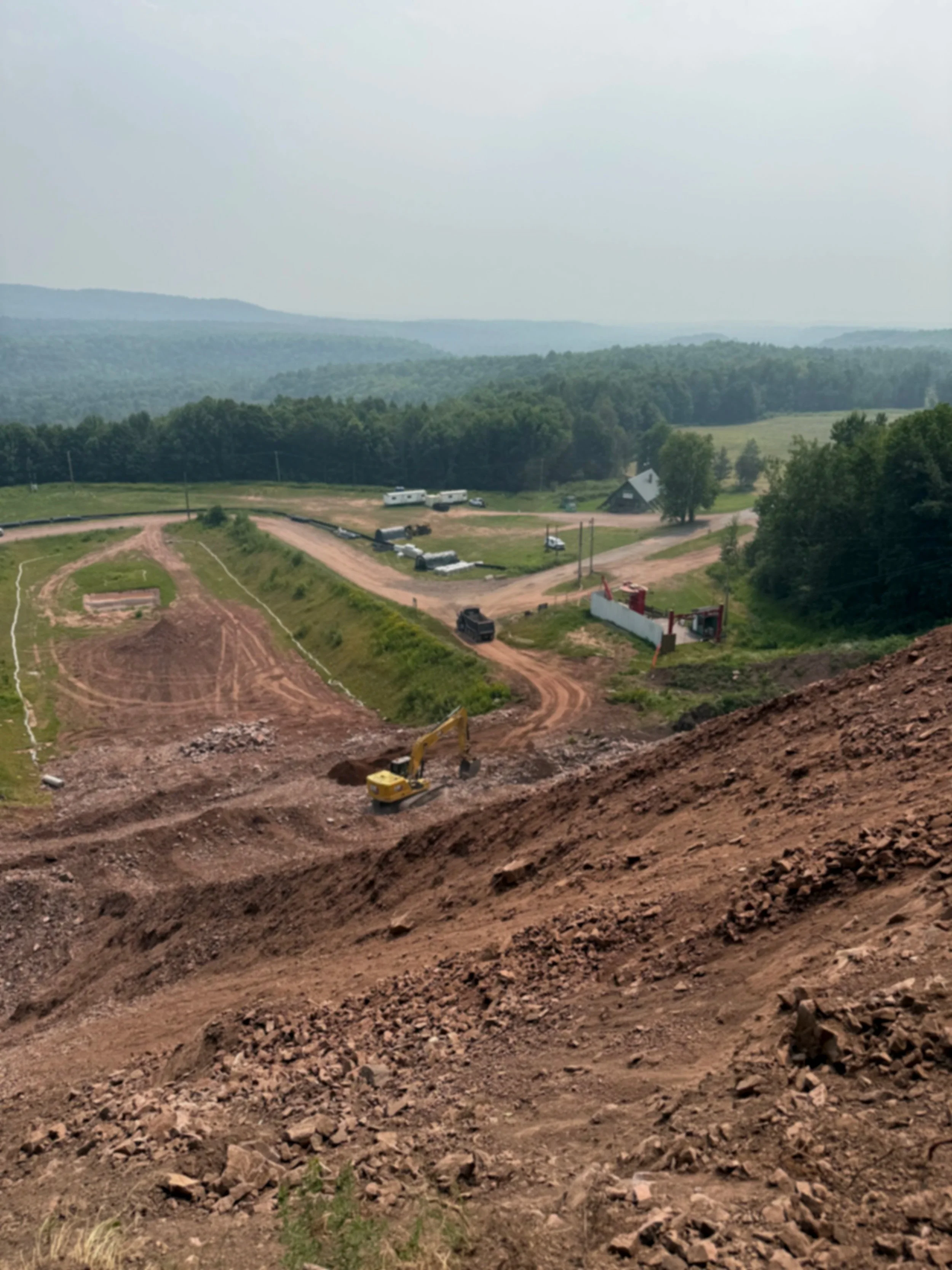 Construction site on a hillside with an excavator at work; dirt and rocks are being moved, and there is a dirt road leading to a lower area. In the background, there are parked cars, trees, a small building, and rolling hills under a cloudy sky.
