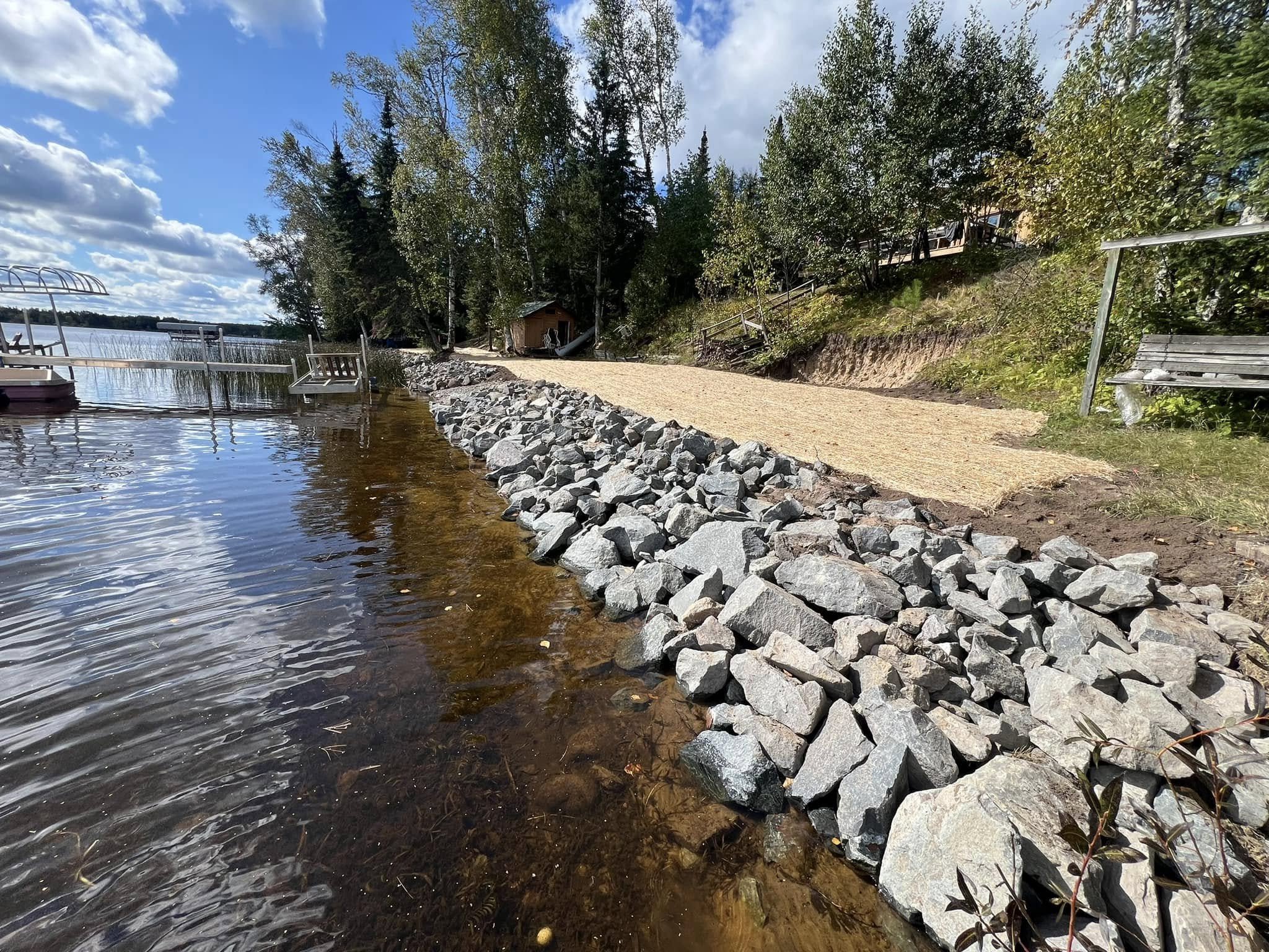A lakeside shoreline with rocks and a sandy path, surrounded by trees, under a partly cloudy sky.