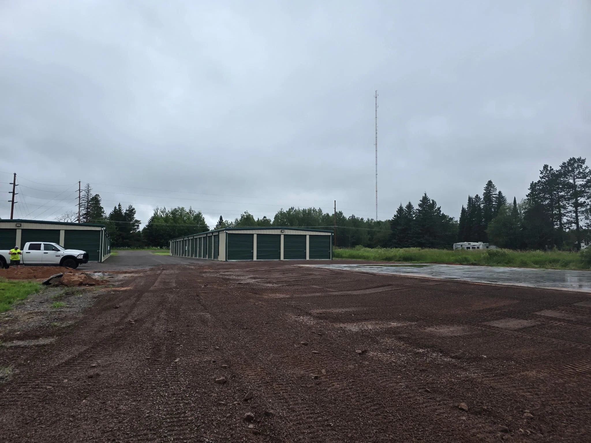An empty lot with freshly laid dirt, a row of storage units or garages, a white pickup truck parked near a person in a yellow safety vest, green trees in the background, overcast sky, and utility poles and a radio tower.