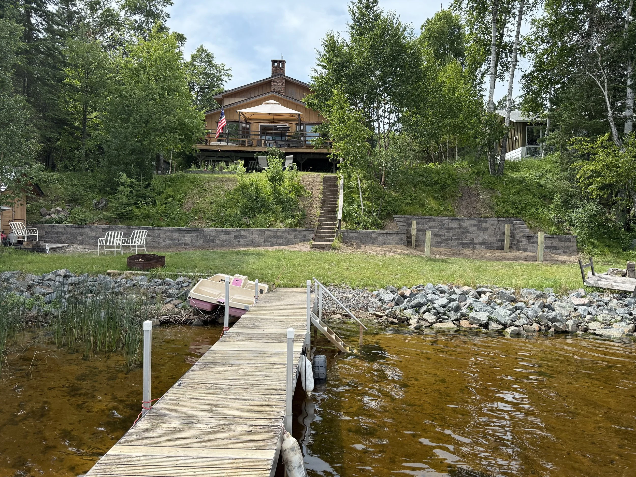 A lakeside house with a deck and an American flag, surrounded by trees, with a staircase leading up from the shore, a dock with a paddle boat, and a fire pit on the lawn.