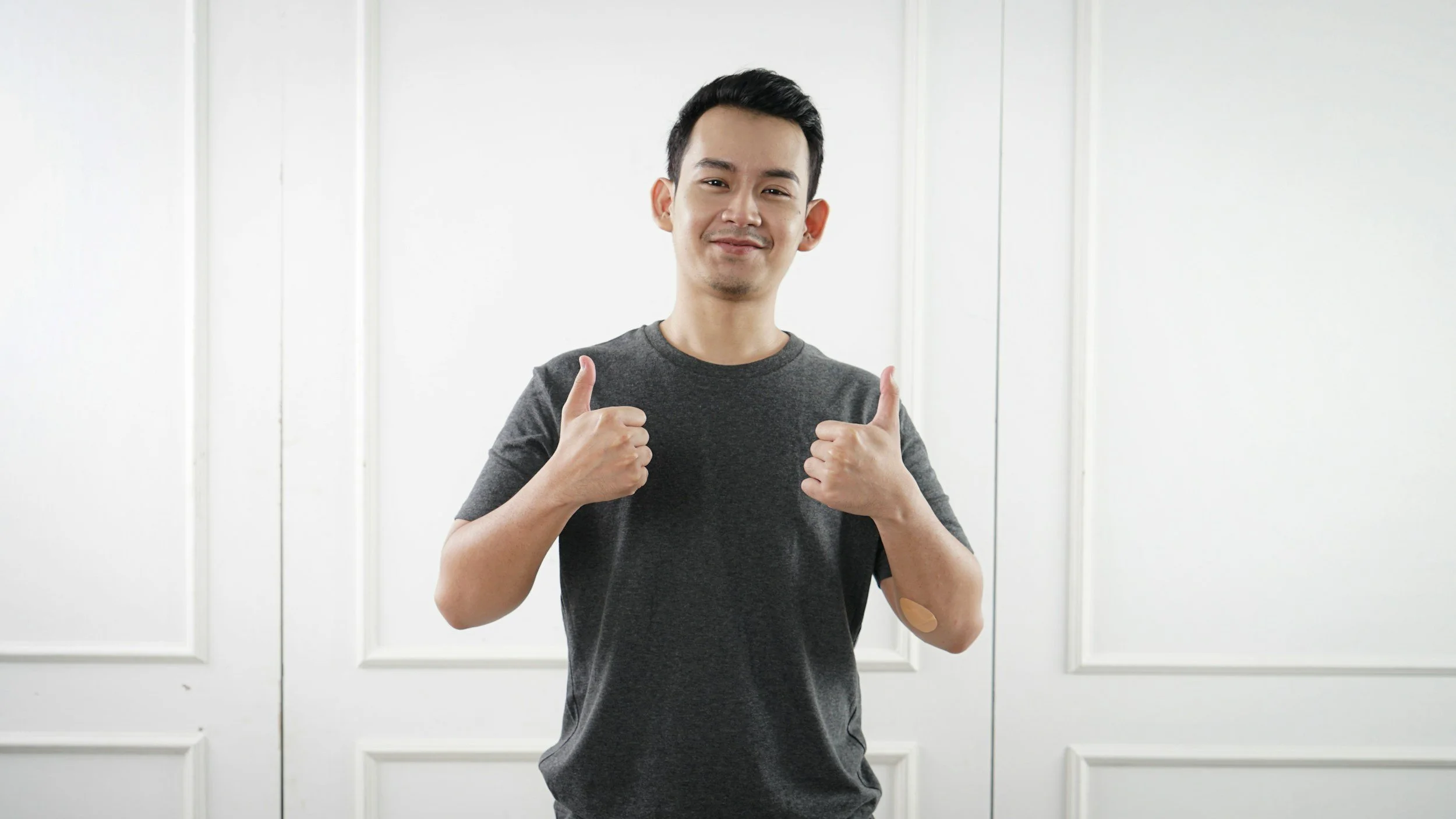 Person in gray t-shirt giving thumbs up in front of white wall.