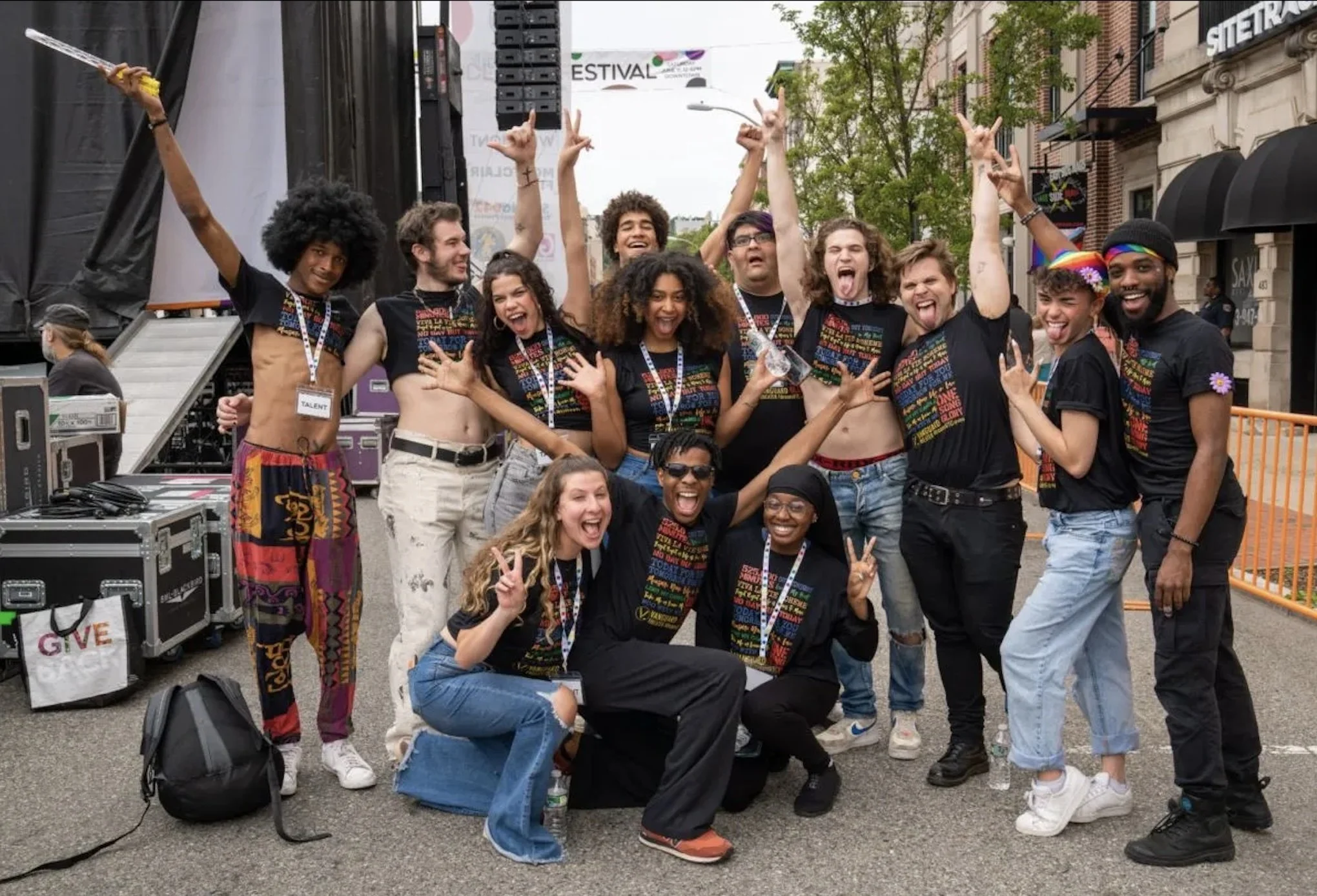 A diverse group of performers from Rent pose together outdoors at Montclair Pride 2022, smiling and cheering with raised arms in front of a stage. They wear matching black T-shirts and appear joyful and celebratory in a city street setting.