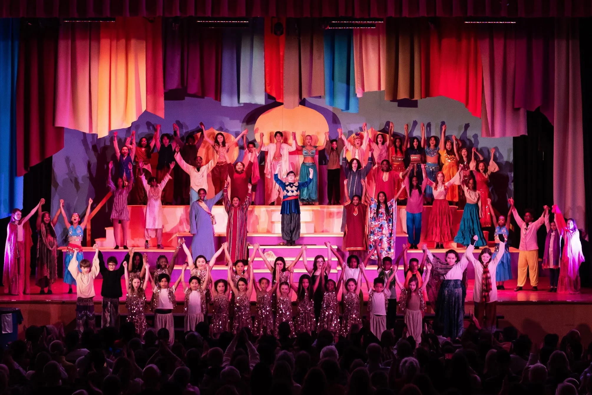 A large cast of children and teens perform a final pose onstage during Aladdin Jr., standing on tiered platforms with arms raised beneath colorful fabric scenery and theatrical lighting.