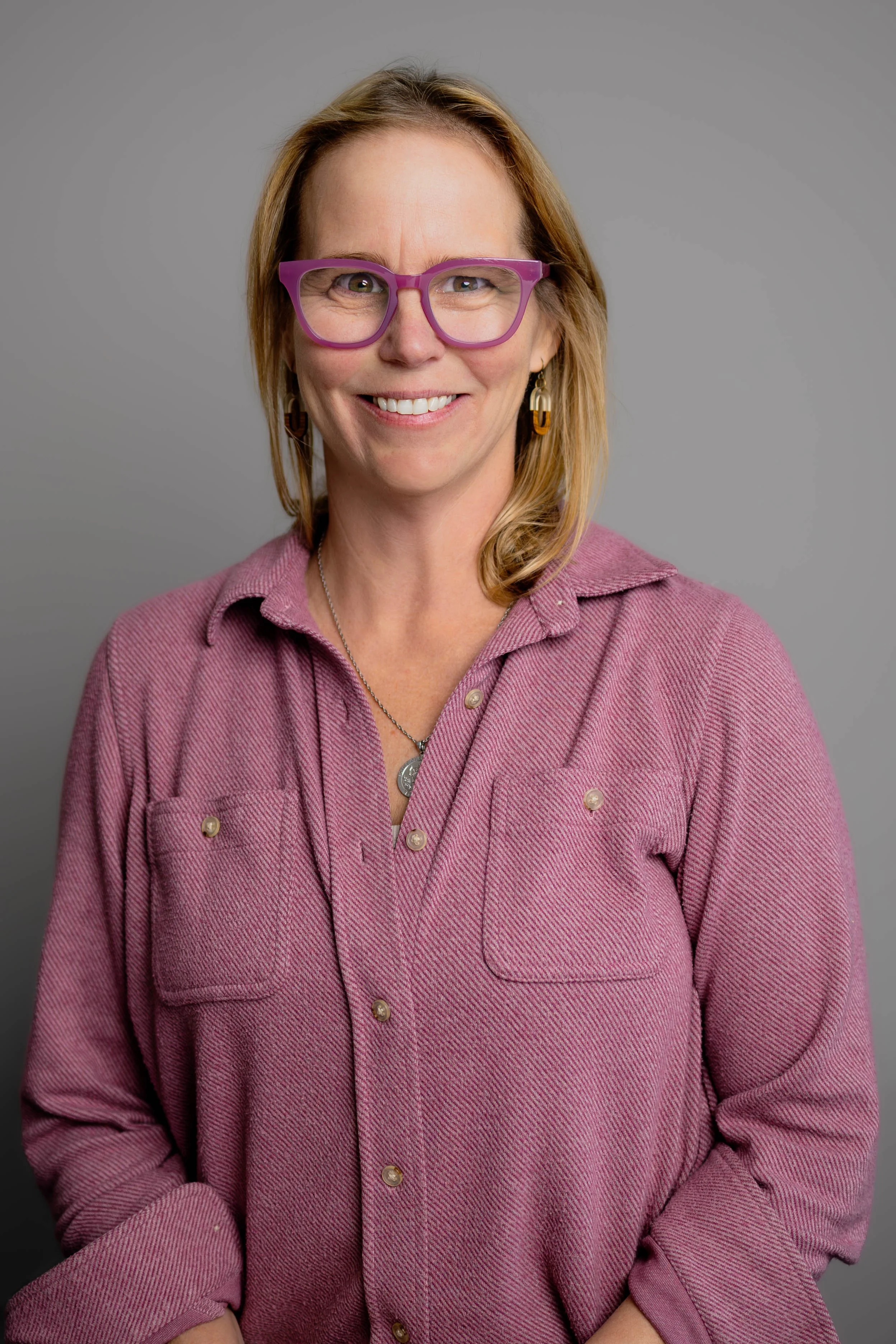 A woman with shoulder-length blonde hair, wearing purple glasses, gold earrings, and a pinkish-purple button-up shirt, smiling at the camera against a gray background.