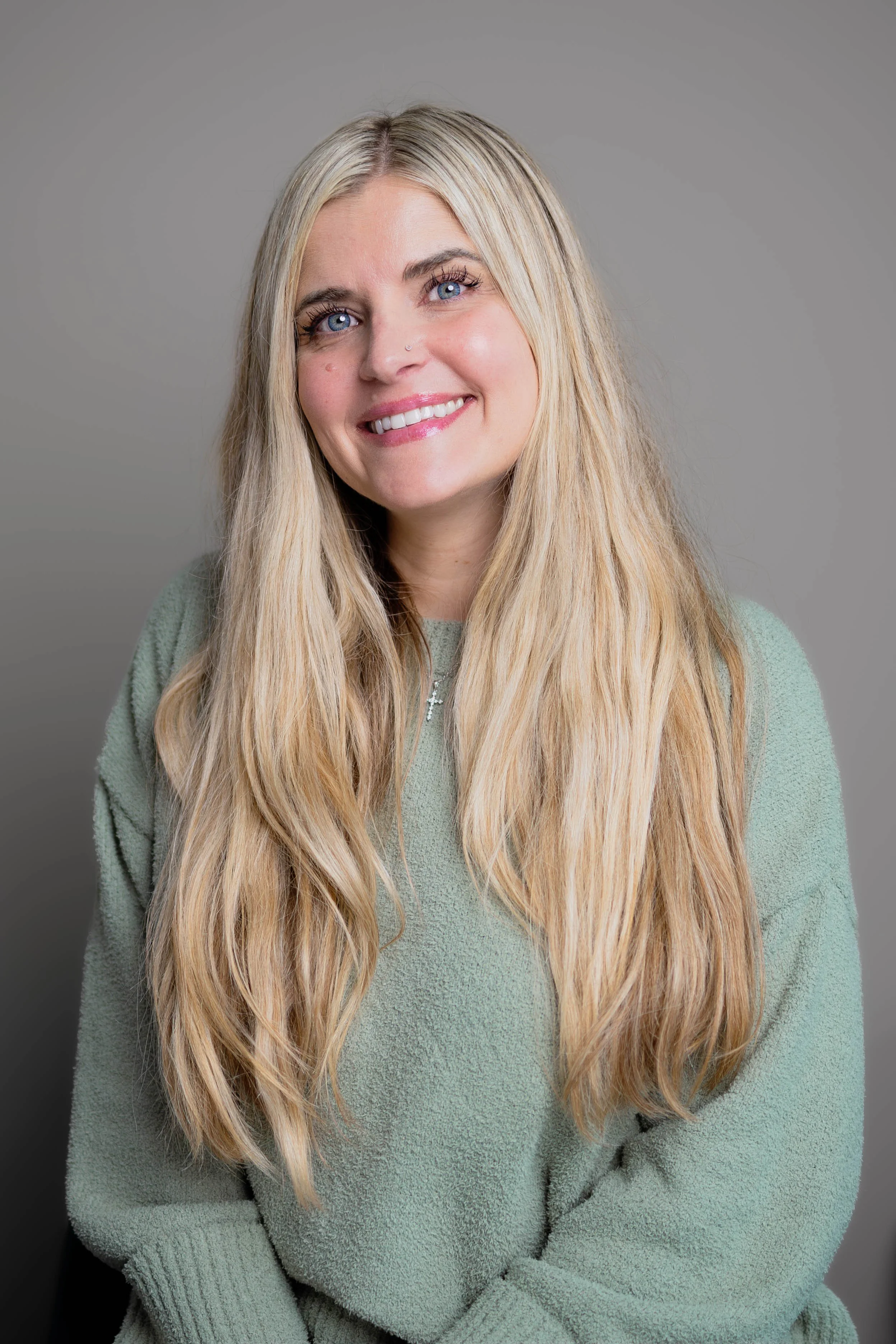 A woman with long blonde hair and blue eyes smiling at the camera, wearing a light green sweater against a gray background.