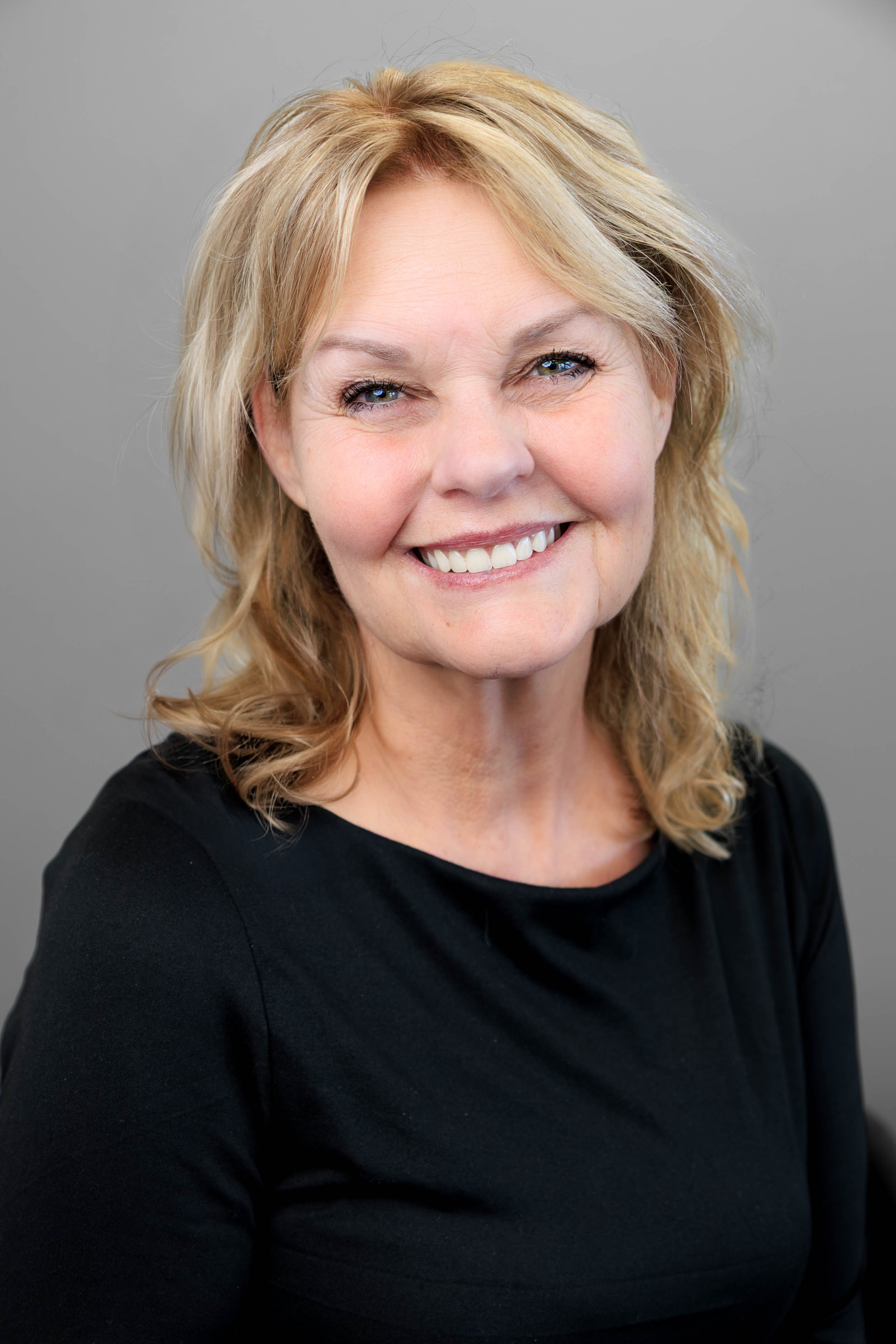 A close-up portrait of a smiling middle-aged woman with blonde hair against a plain gray background.