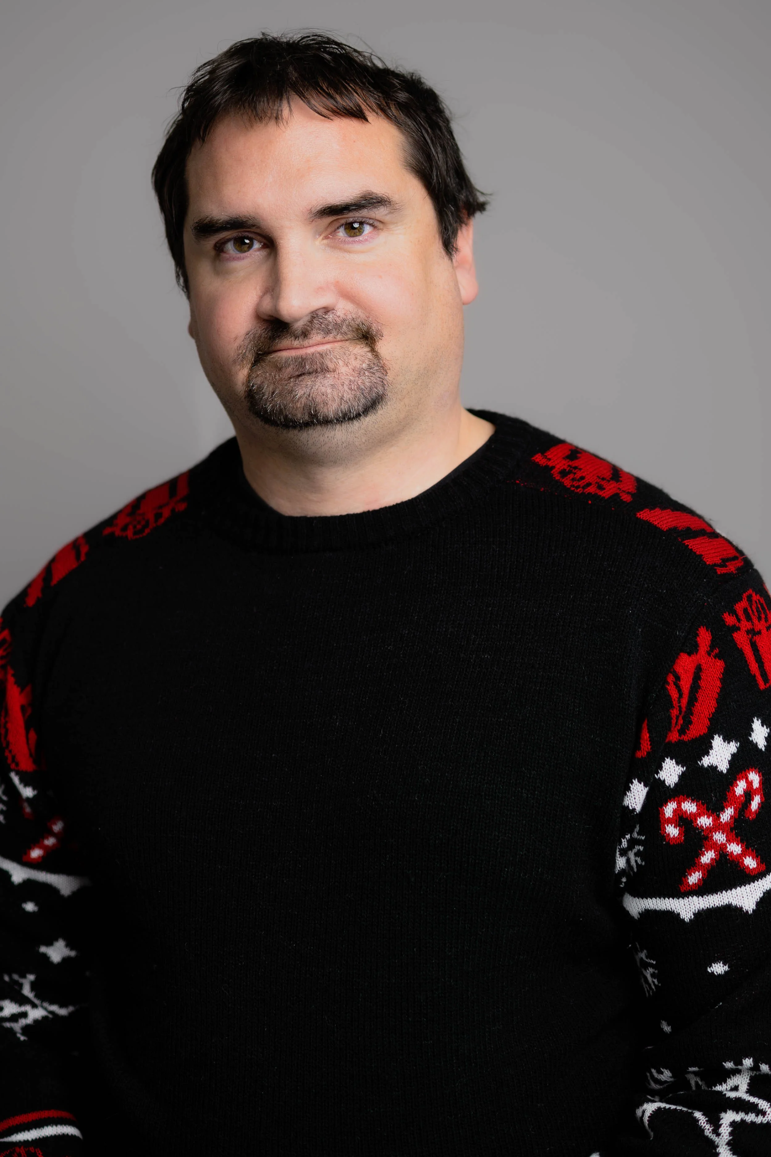 Portrait of a man with dark hair and a goatee, wearing a black sweater with red and white Christmas-themed patterns on the sleeves, standing against a plain gray background.