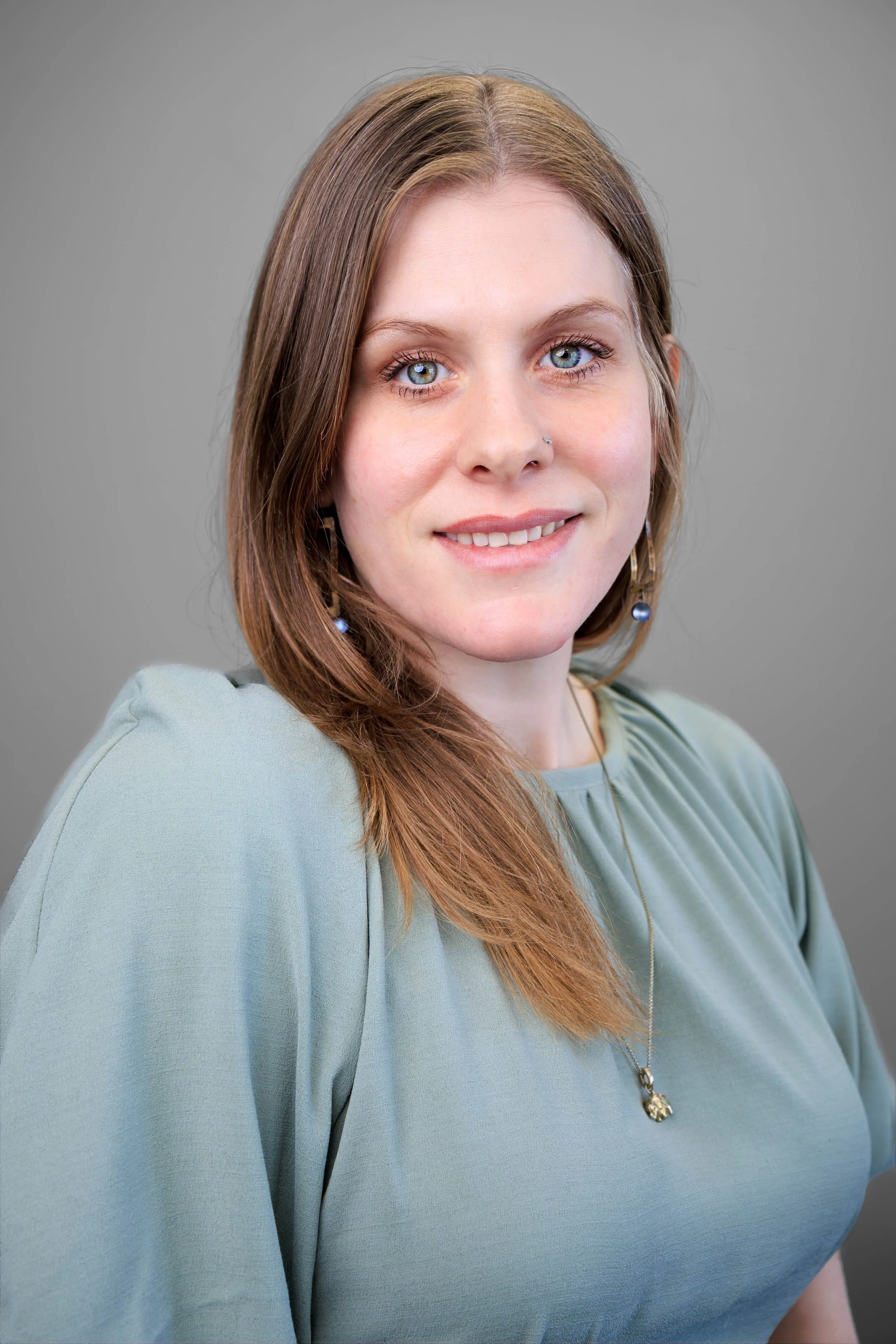 Portrait of a woman with light skin, blue eyes, long light brown hair, wearing a grayish-green top, gold necklace, and earrings with blue beads, smiling against a gray background.