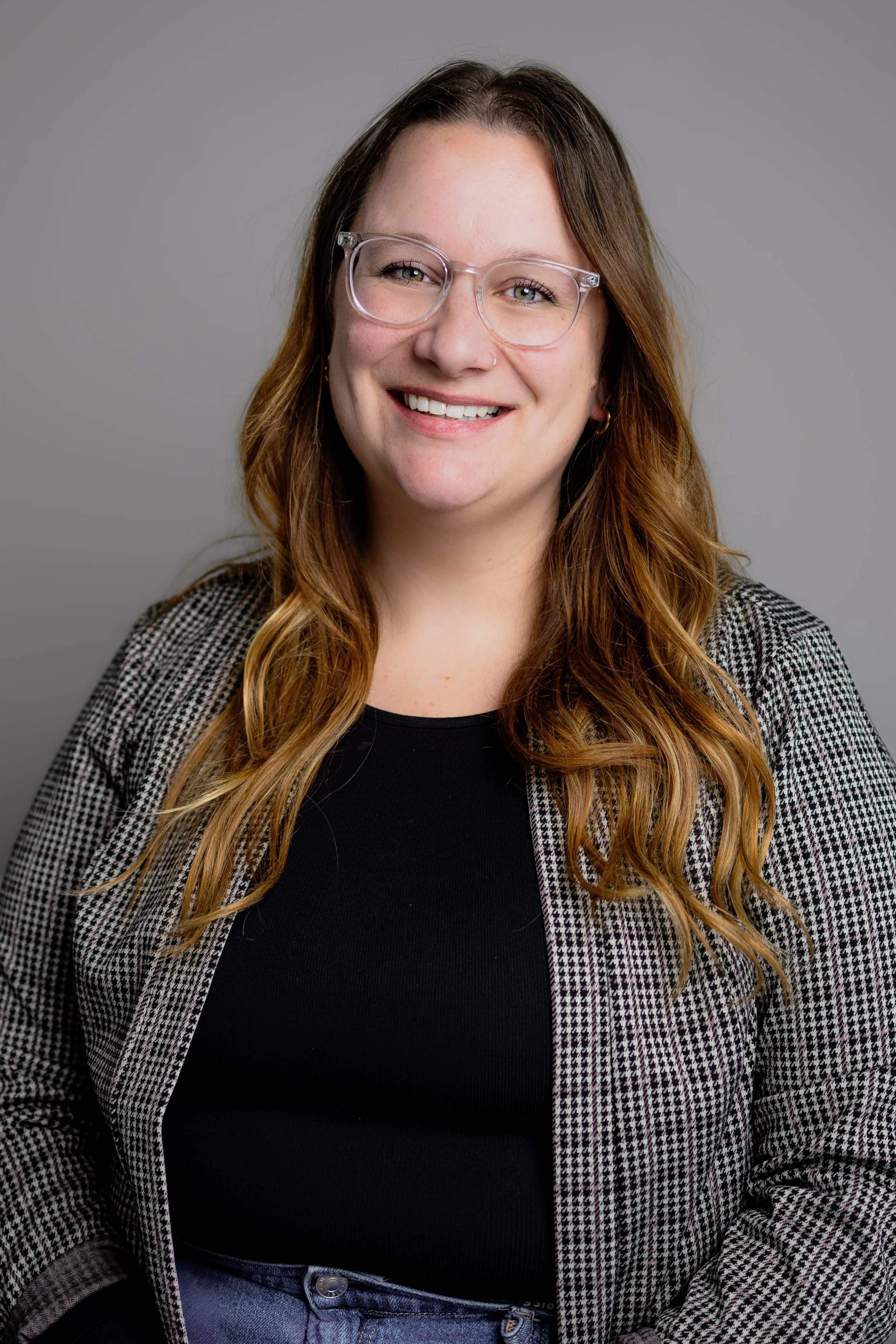 A woman with long, wavy, reddish-brown hair and glasses smiling against a gray background.