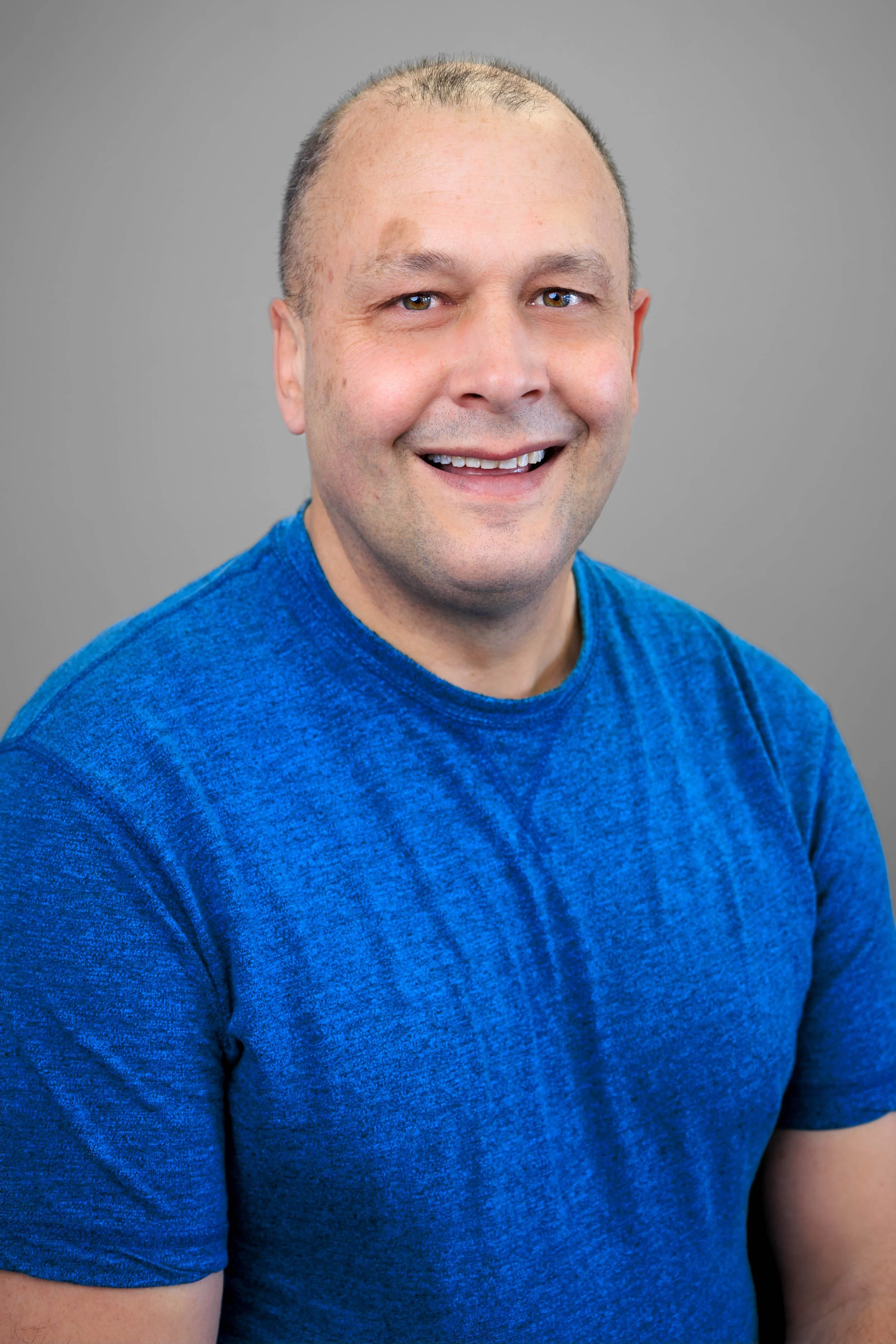 A smiling man with short hair wearing a blue t-shirt, posing against a plain gray background.