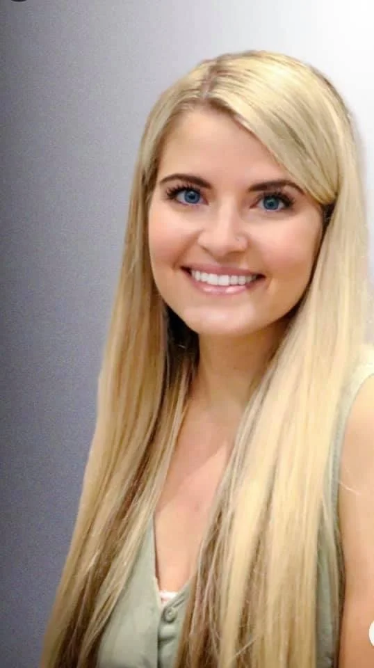 Close-up of a smiling young woman with long blonde hair and blue eyes, wearing a light-colored top, standing against a plain background.