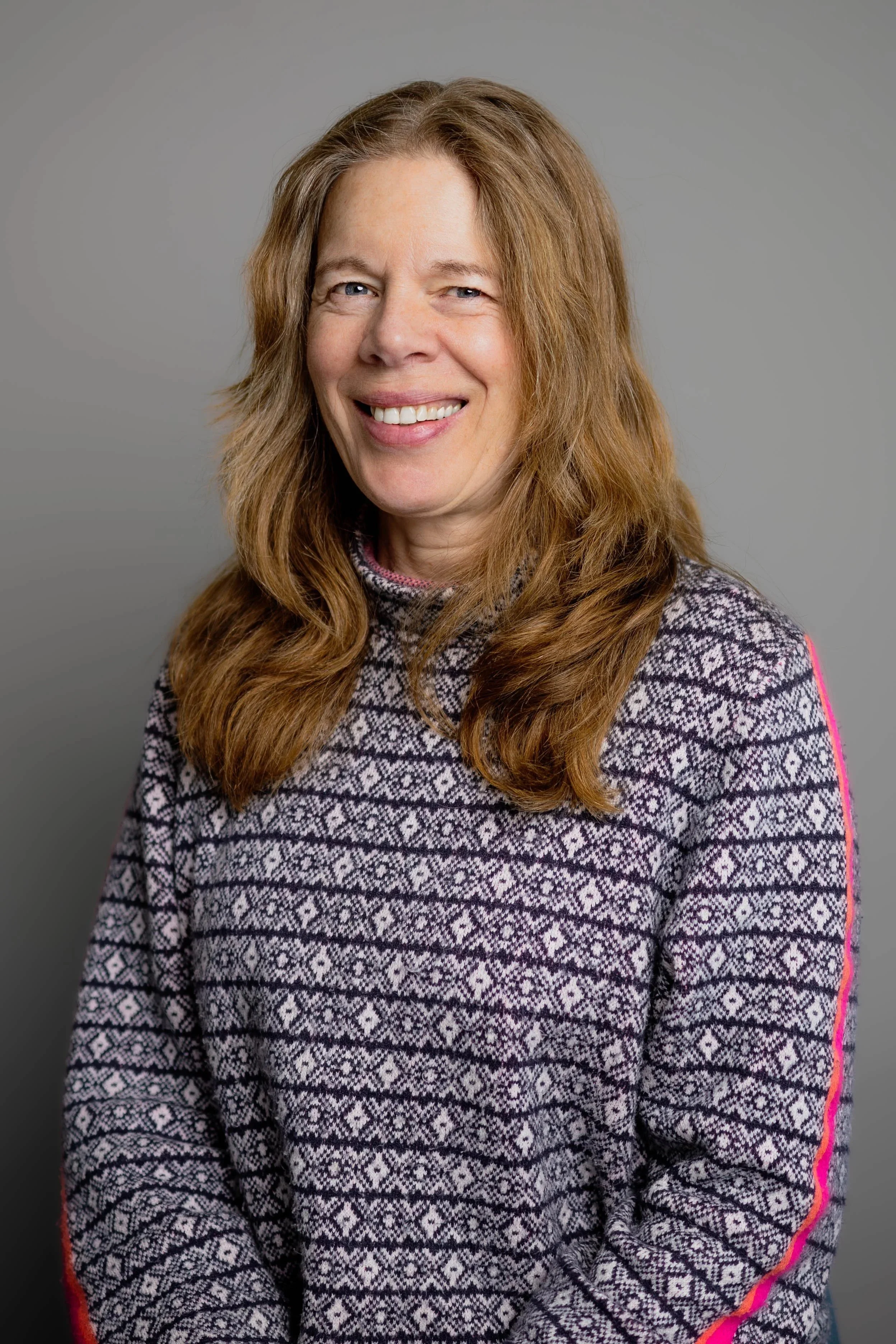 Portrait of a woman with long, wavy red hair, smiling, wearing a patterned sweater with pink accents on the sleeves, against a neutral gray background.