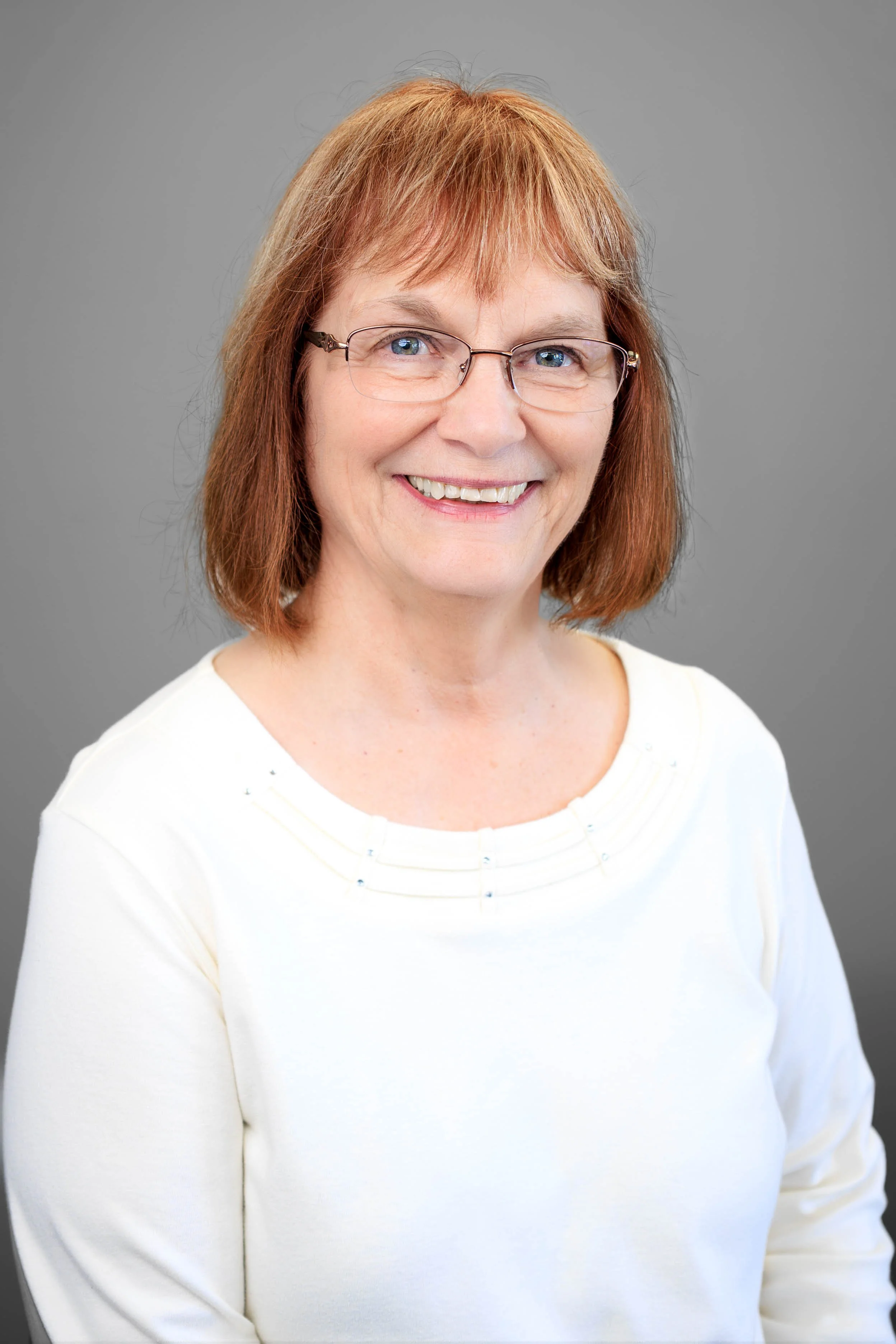 Portrait of a middle-aged woman with reddish-brown hair, glasses, smiling, wearing a white top, against a gray background.