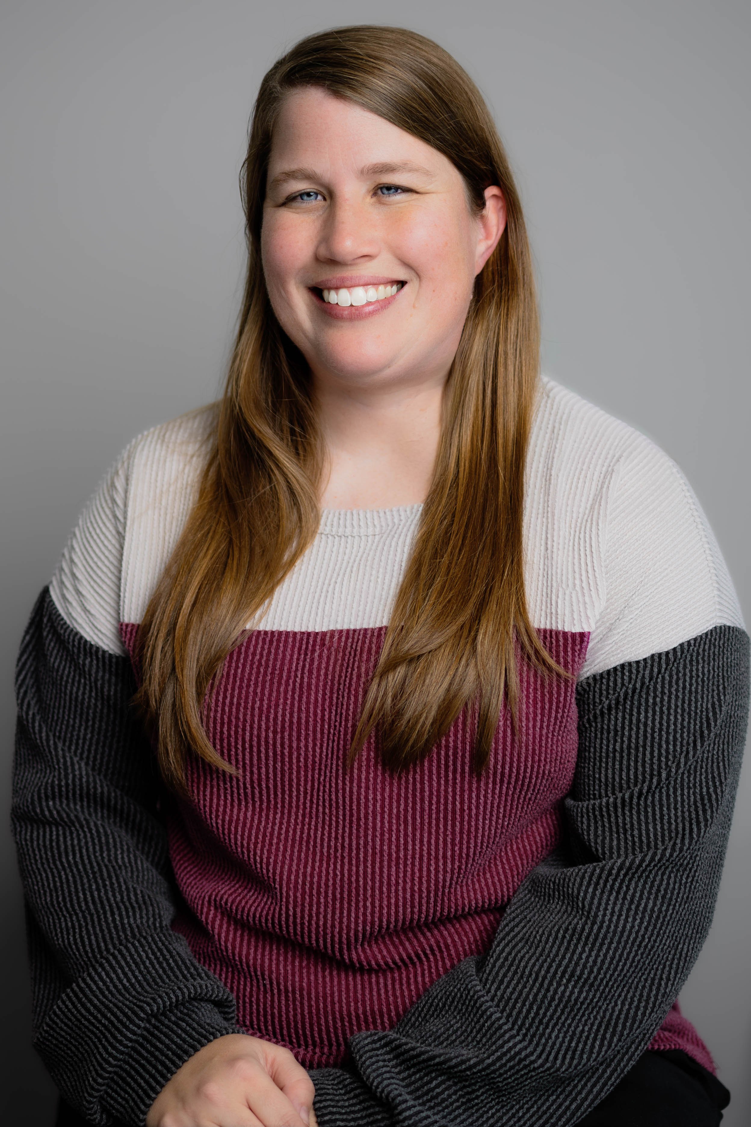 A woman with long, straight, reddish-brown hair smiling at the camera, wearing a color-blocked sweater with white, maroon, and black sections, against a plain gray background.
