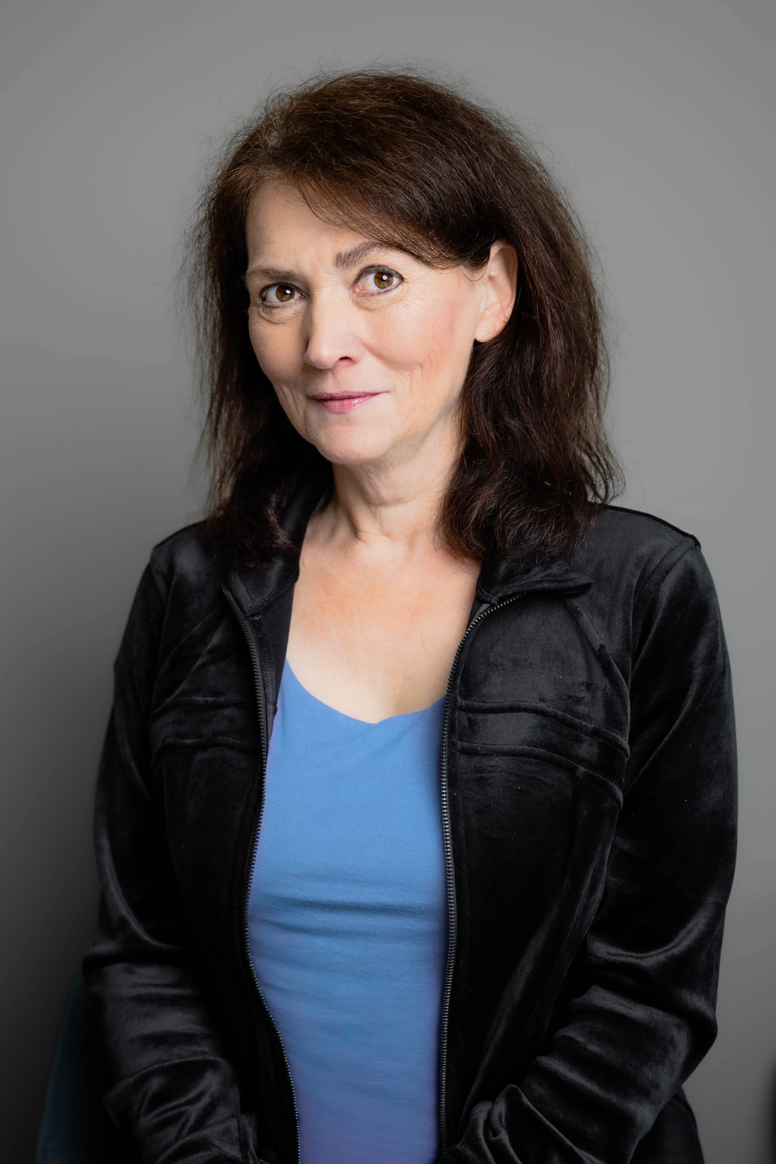 A portrait of an older woman with dark brown hair, wearing a black jacket over a blue top, against a plain gray background.