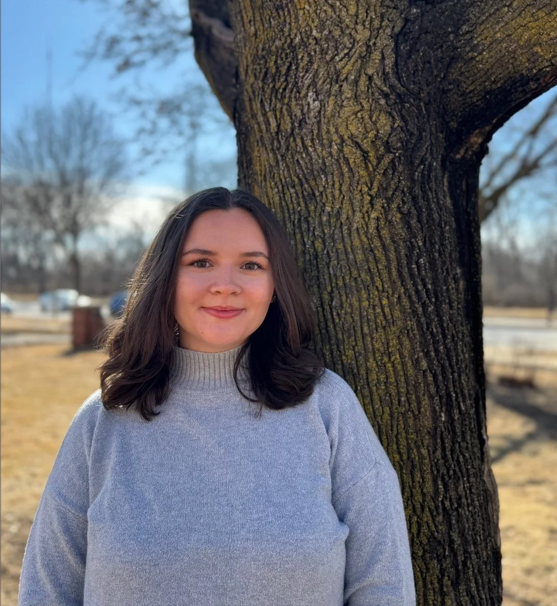 A young woman with shoulder-length dark hair, wearing a light gray sweater, stands outdoors beside a large tree with textured bark. In the background, there are some leafless trees, a grassy area, and a clear blue sky.