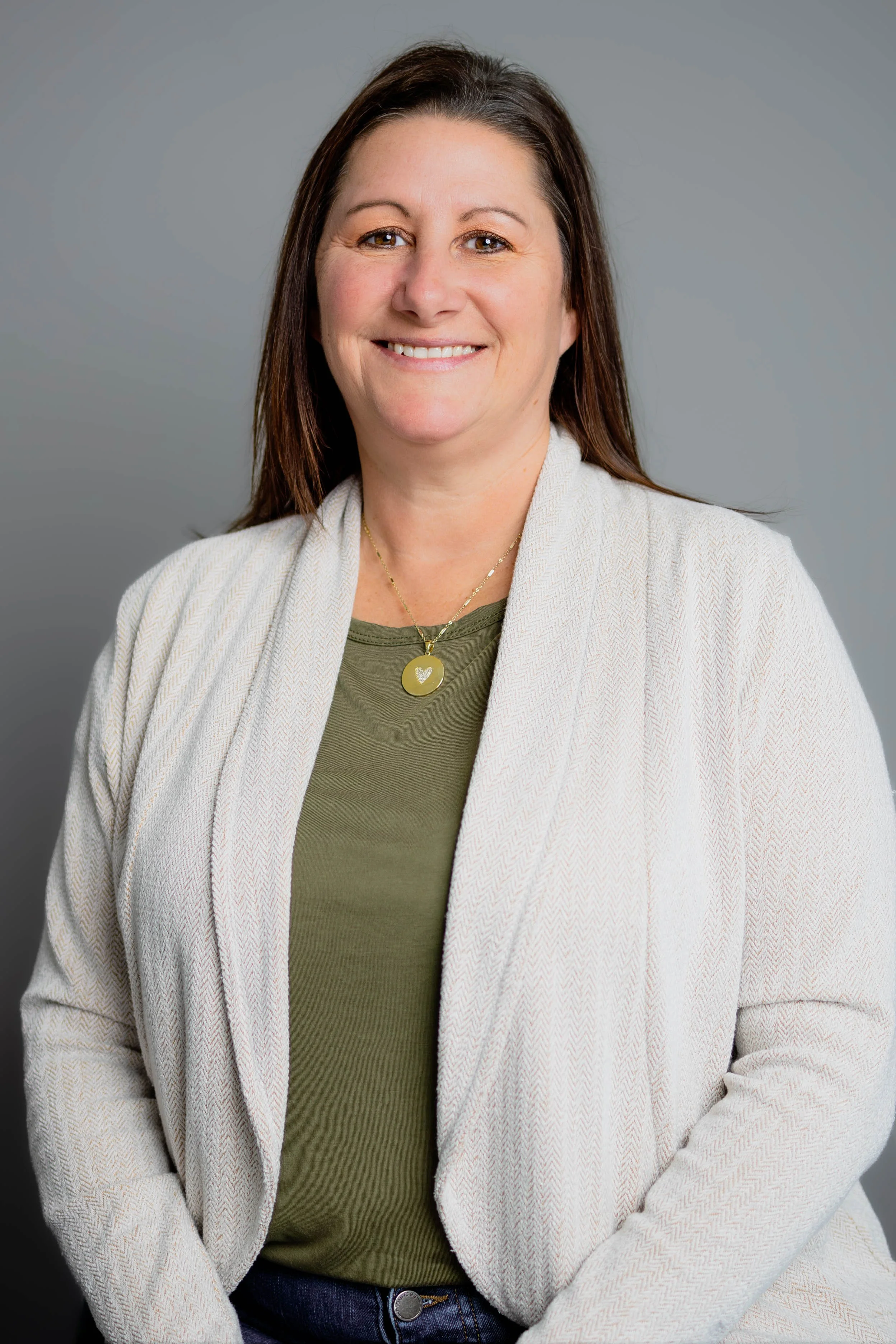 Portrait of a woman with long brown hair, wearing a white blazer, a green top, and a gold necklace with a heart pendant, smiling against a gray background.