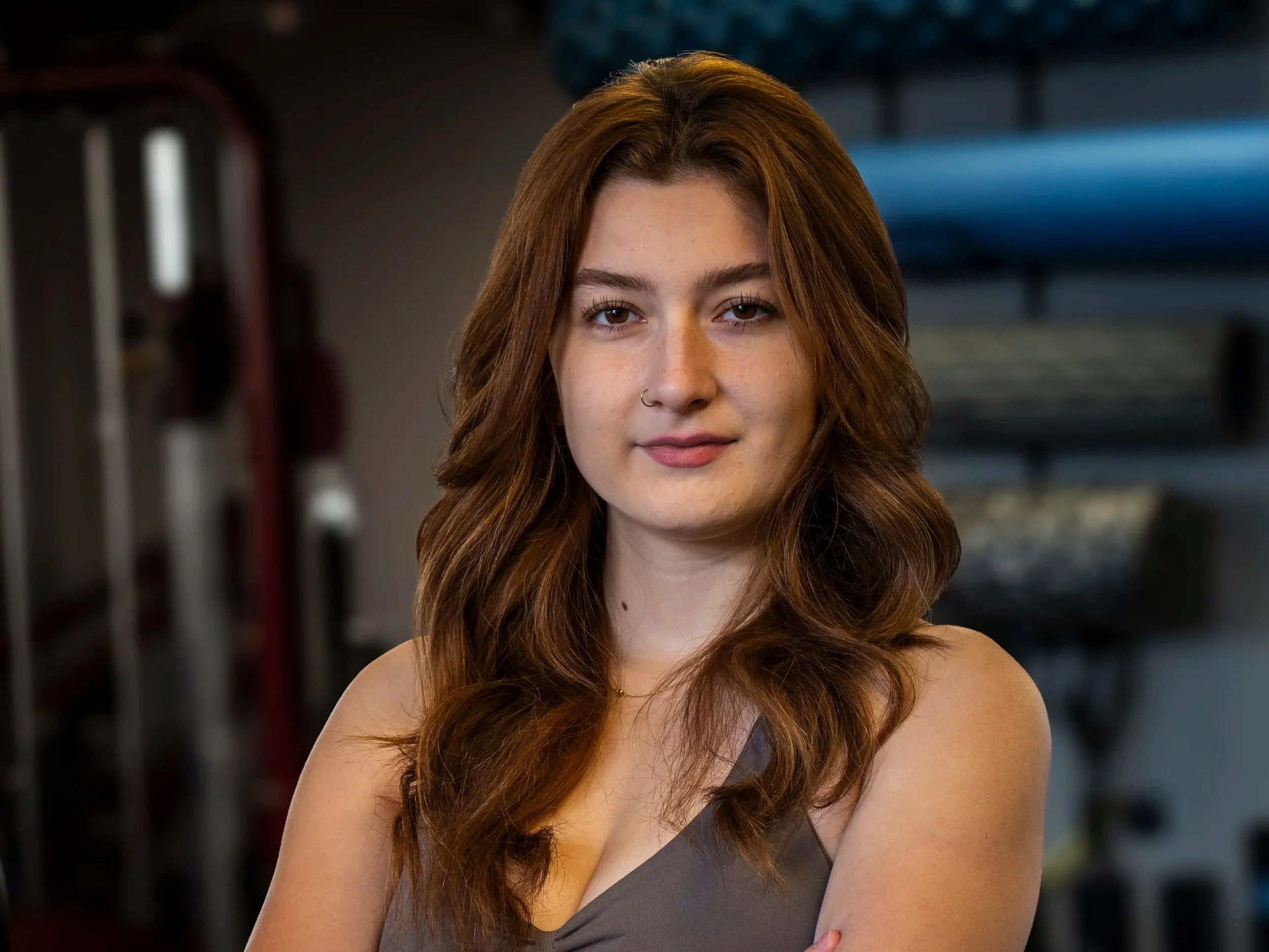 A young woman with long wavy brown hair standing with arms crossed inside a gym with exercise mats and equipment in the background.