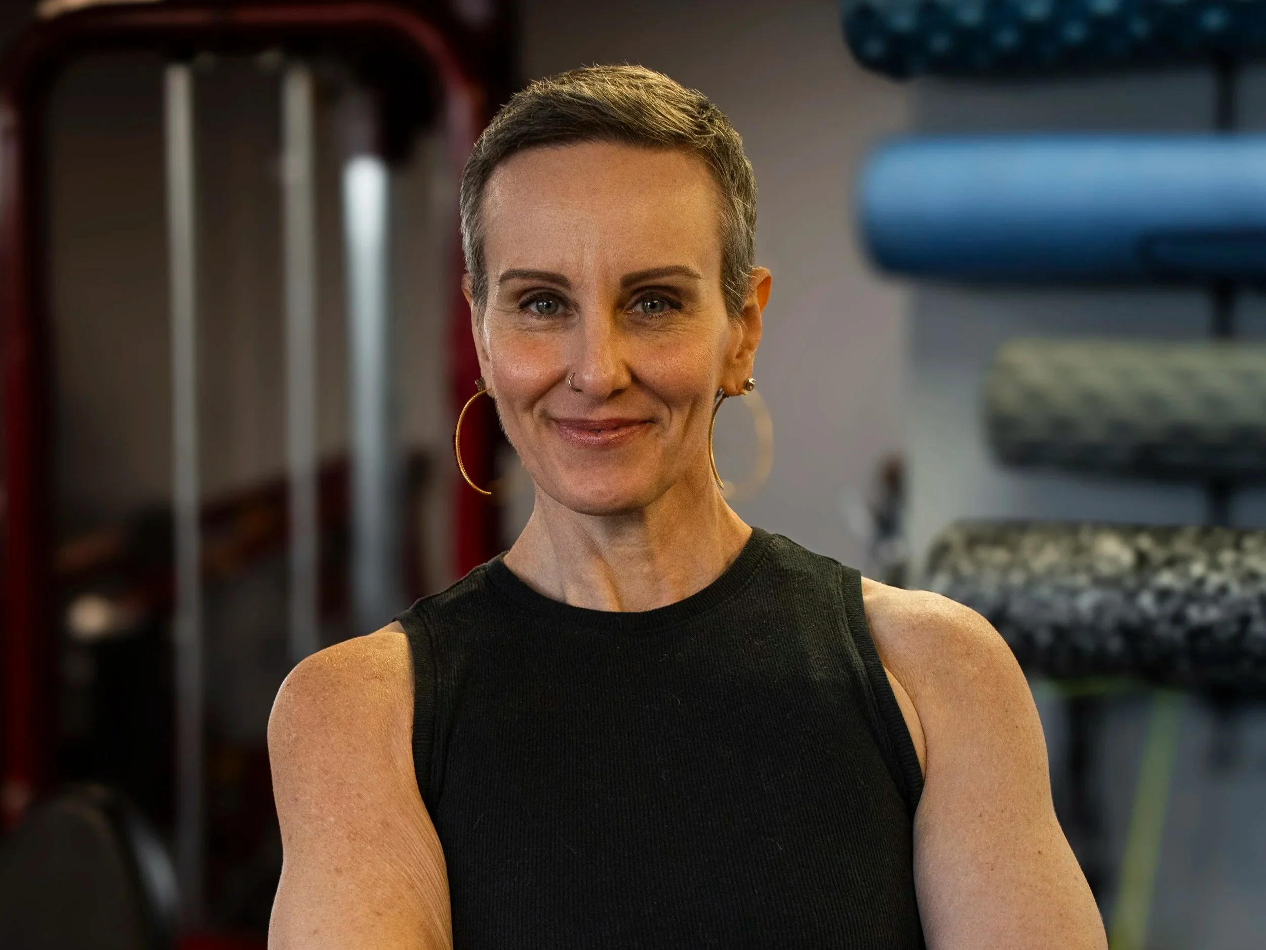 A confident woman with short hair, wearing a black athletic top and hoop earrings, standing with arms crossed in a gym with workout equipment in the background.