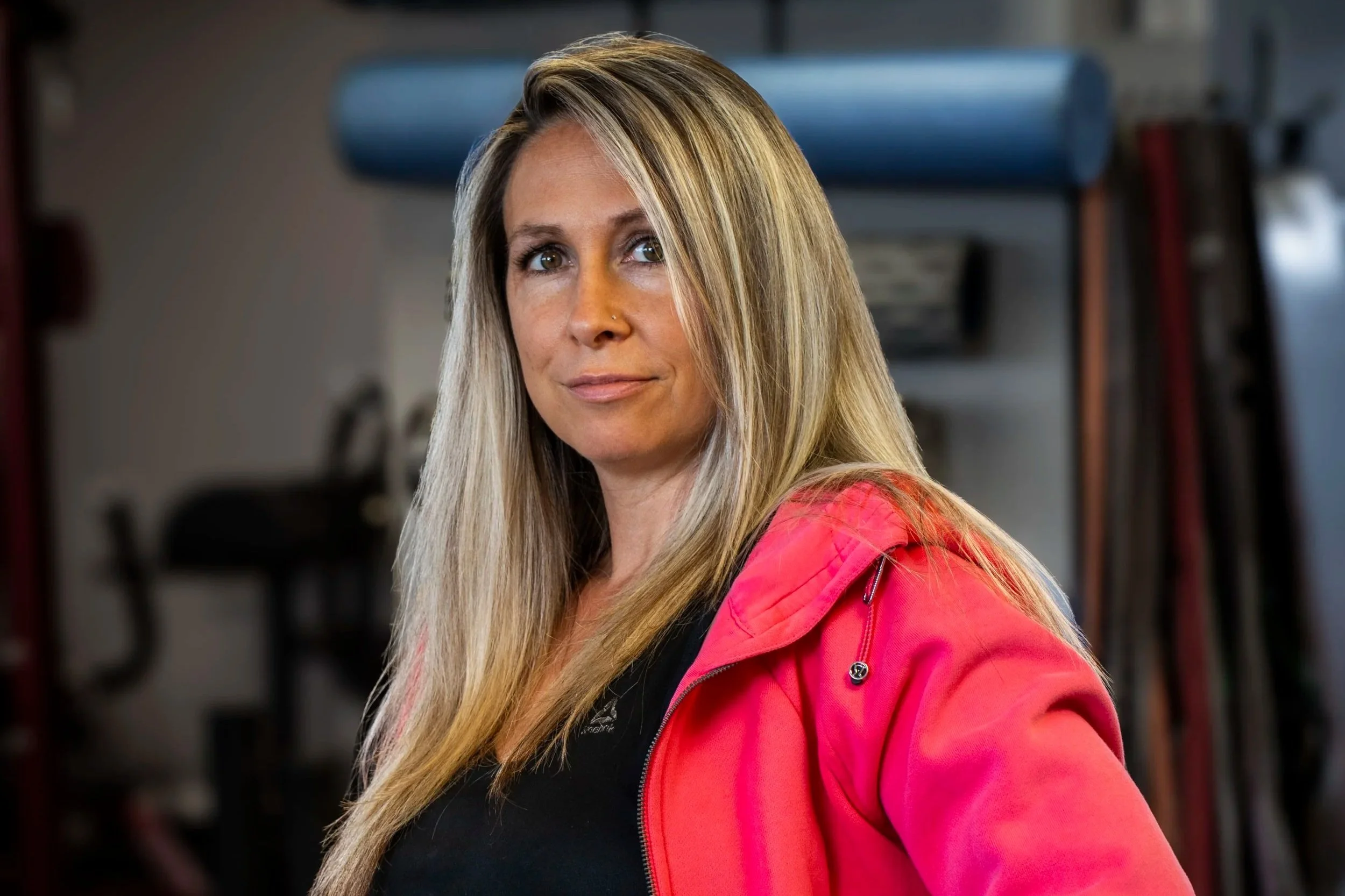 A smiling woman with long, blonde hair and a nose piercing in a gym, with equipment and weights in the background.