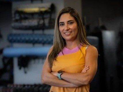 Woman wearing a yellow workout top and dark workout pants, standing with arms crossed in a gym with workout equipment in the background.