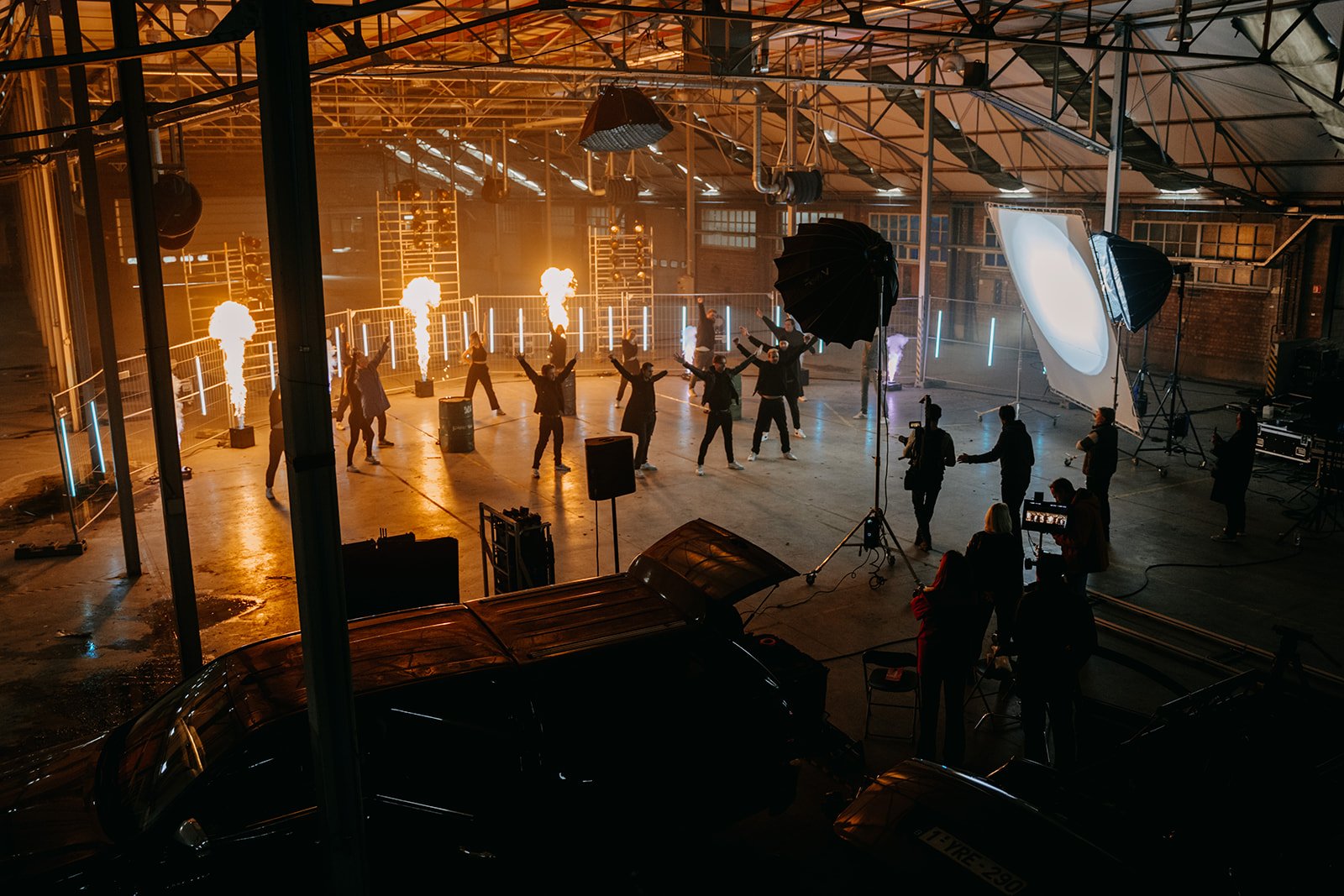 Opname van een muziekvideo in een grote industriële ruimte met mensen die dansen en lichteffecten van vuur en fotoverlichting.