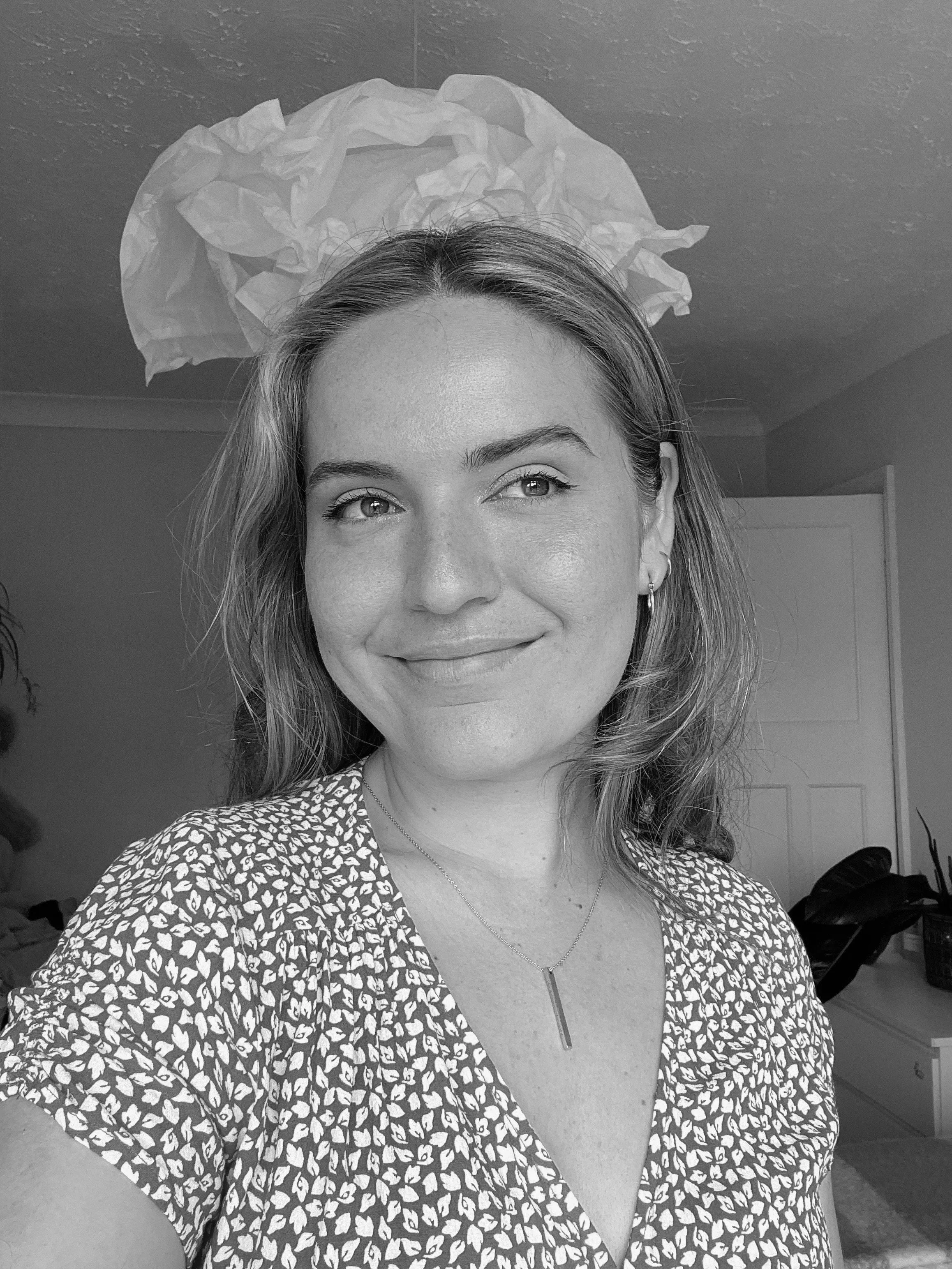 A woman taking a selfie indoors, smiling and looking to the side, with a crumpled paper ball hanging from the ceiling behind her.
