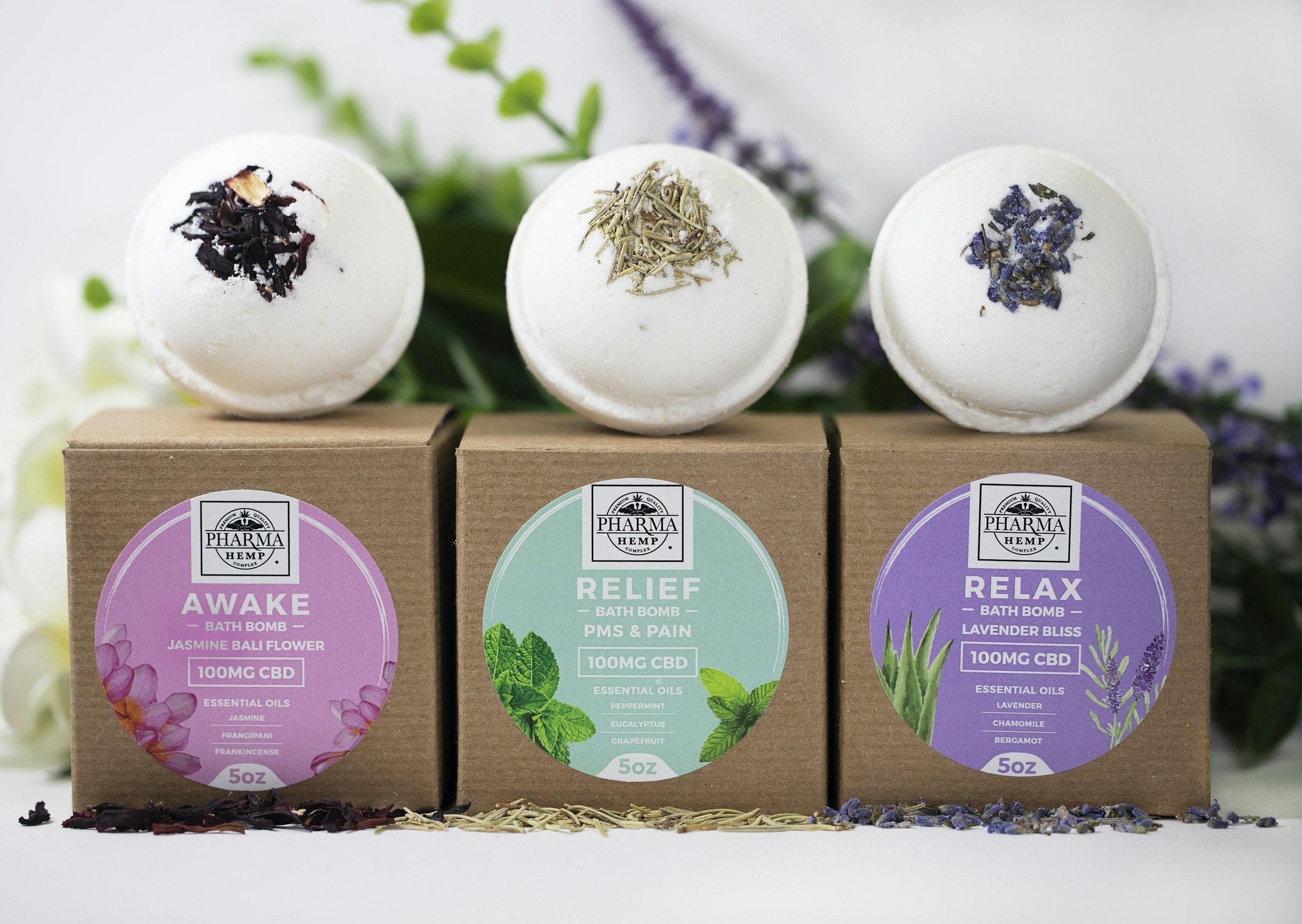 Three boxes of CBD bath bombs labeled Awake, Relief, and Relax, each with a different essential oil topping, and three bath bombs on top with dried herbs or flowers, set against a backdrop of flowers and greenery.