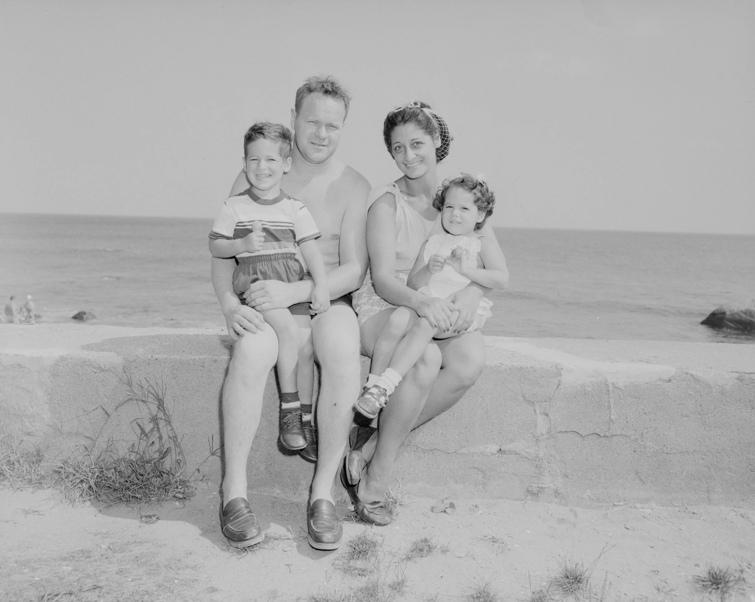 A black and white photo of a family of four sitting on a concrete wall at the beach. The family includes a man, woman, and two young children, with the ocean in the background.