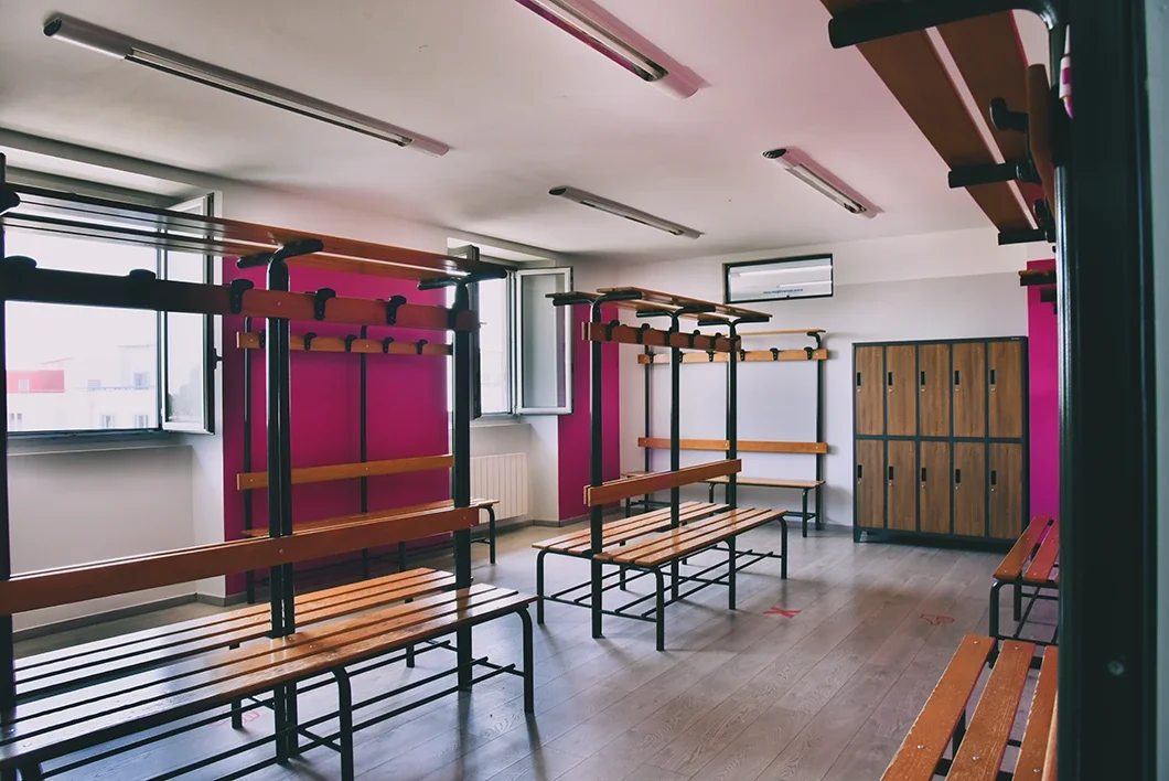 Spogliatoio scolastico con panche e armadietti di legno, pareti colorate in rosa e bianco, finestre aperte, con pavimento in legno.