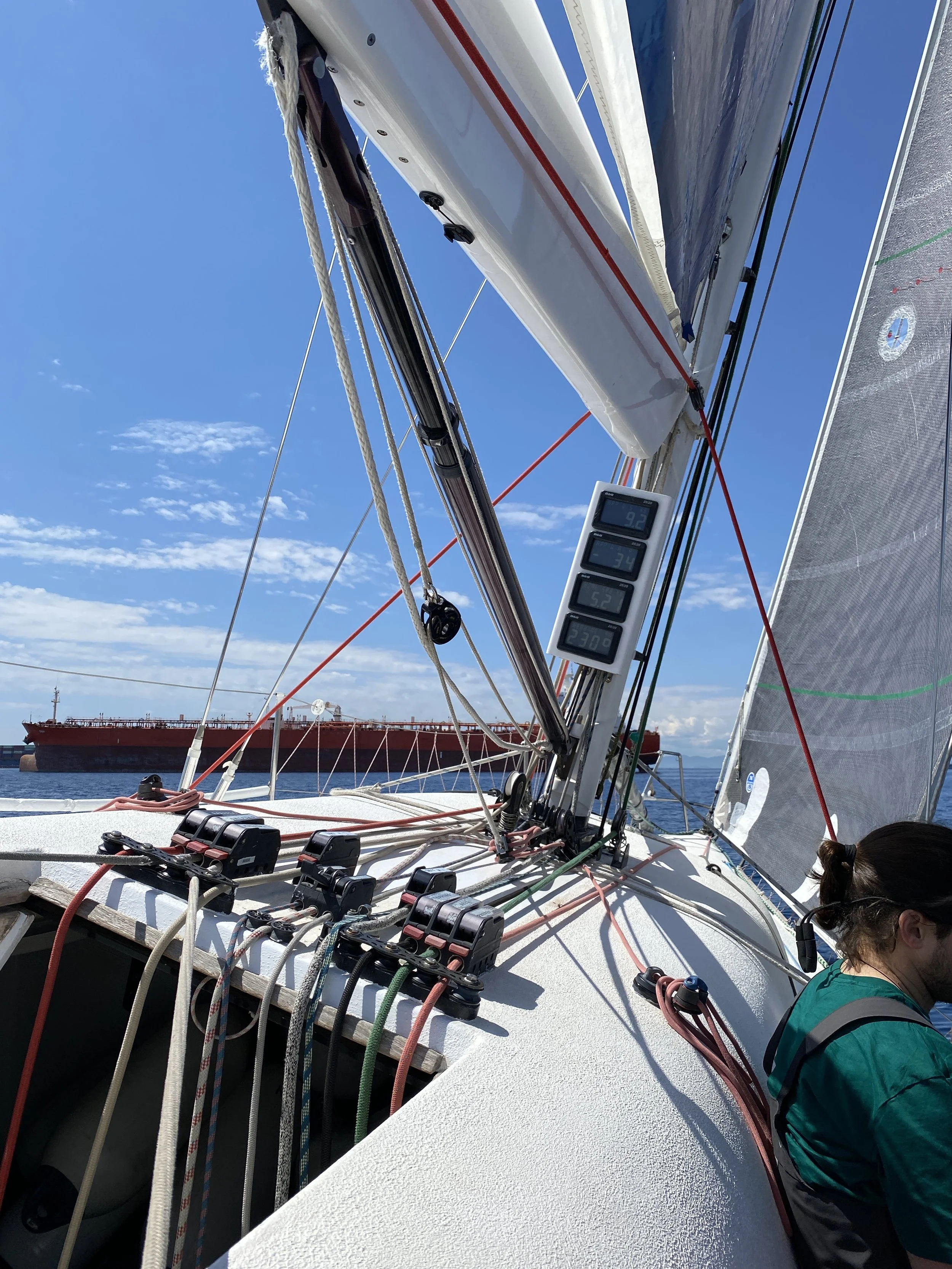View from a sailboat showing its rigging, sails, and instruments, with a large cargo ship in the background on the water under a clear blue sky.