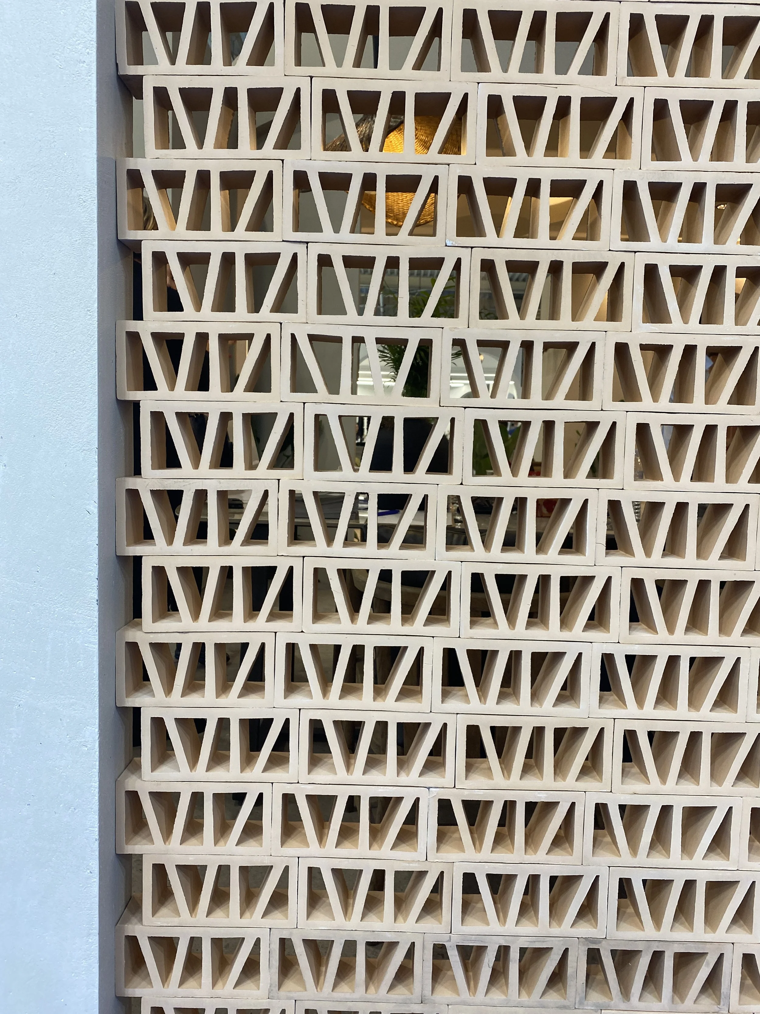 A wall with a decorative white grid pattern, revealing a yellow lamp and some indoor plants behind it.