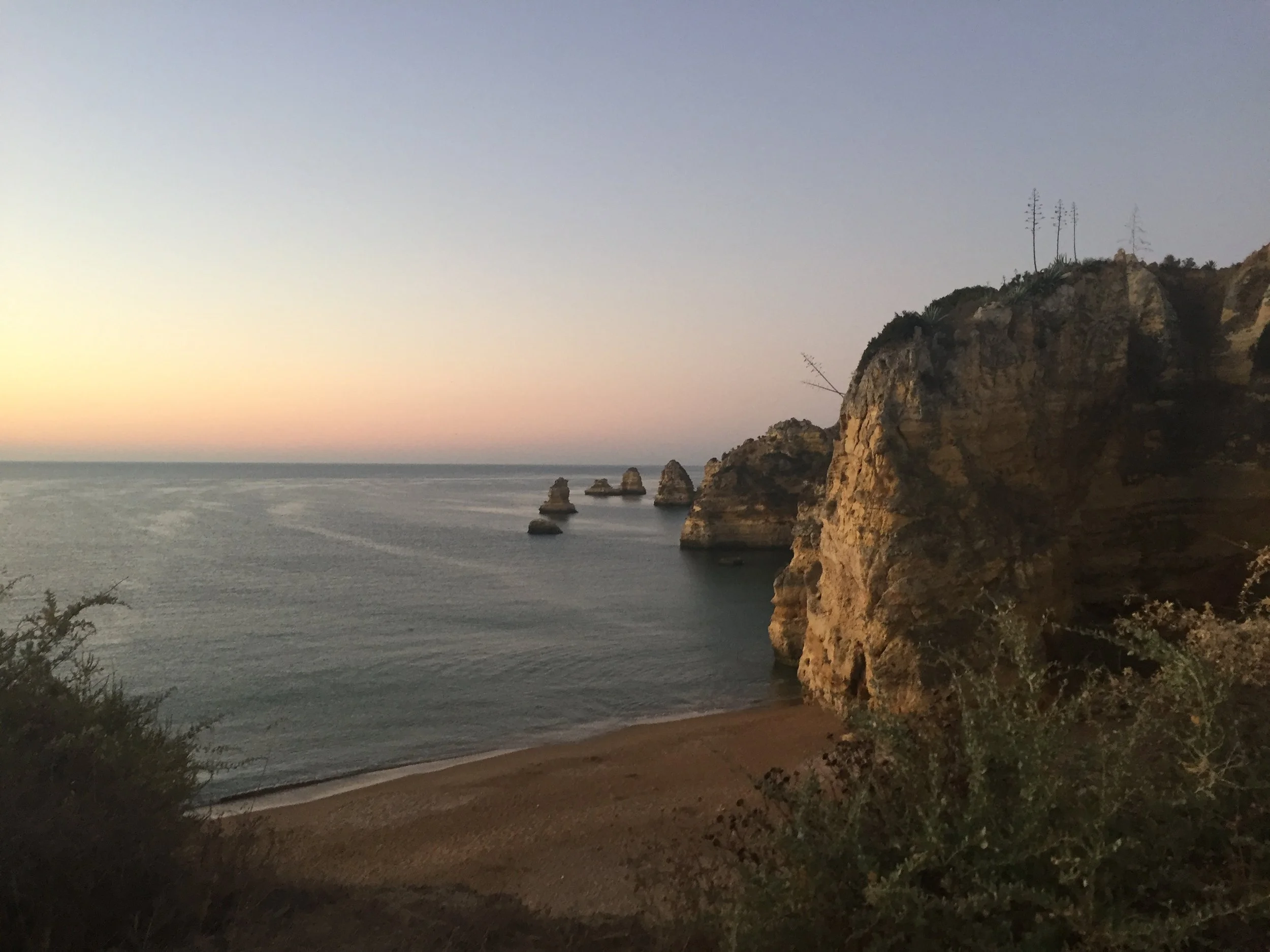 A coastal scene at sunset with a rocky cliff on the right side, sparse trees on top, and the ocean in the background under a clear sky.