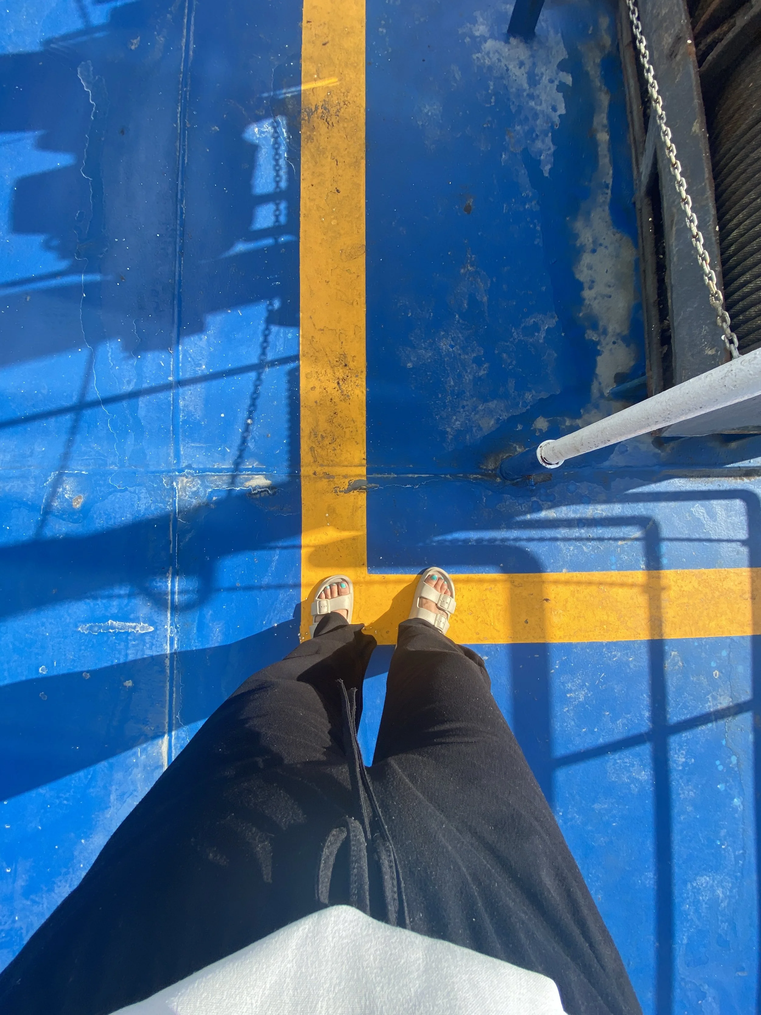 A person standing on a blue platform with yellow safety lines, wearing sandals and black pants, with shadows and reflections visible on the surface.