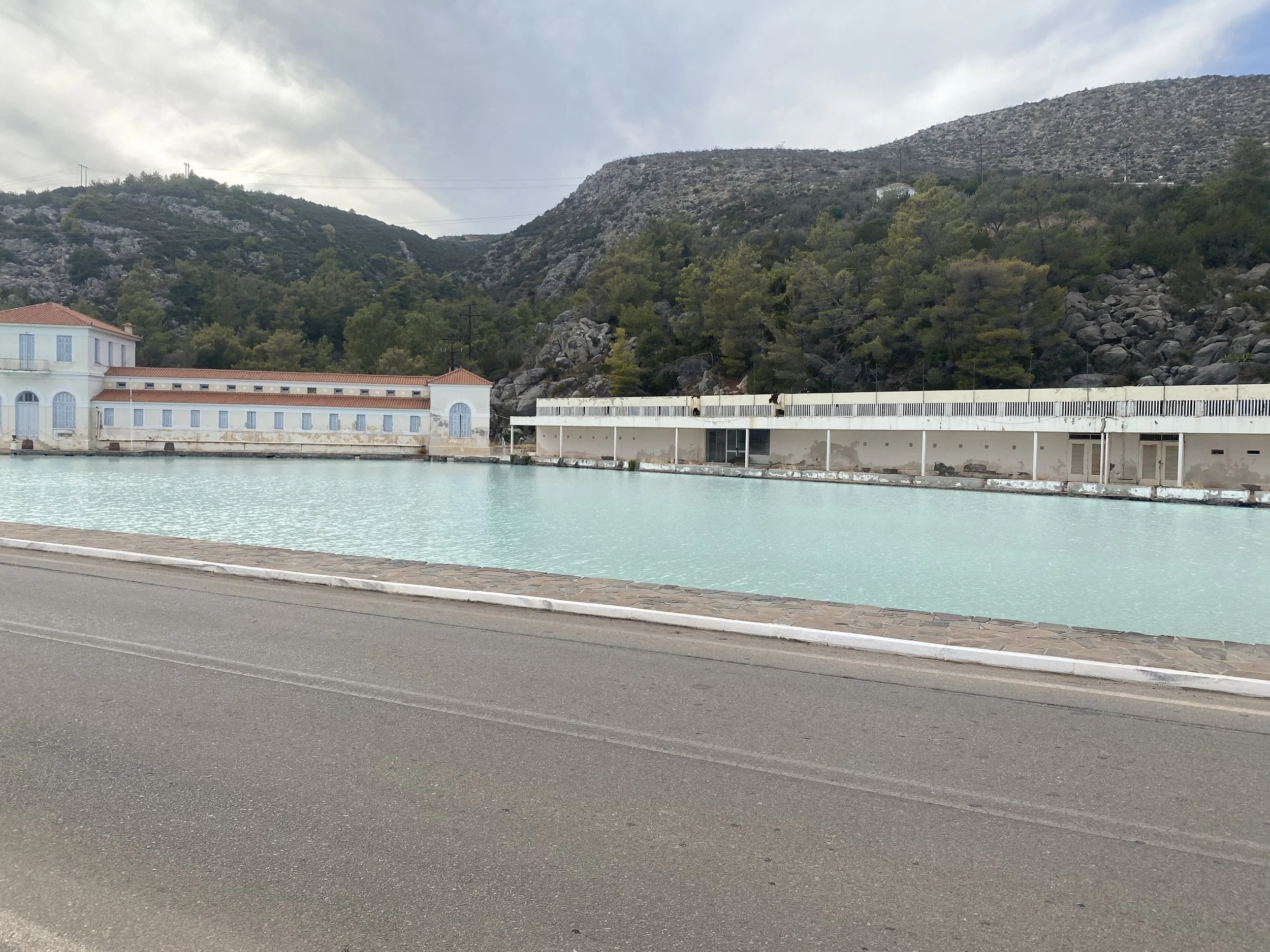 A view of a turquoise lake or pond with a paved road in the foreground, surrounded by hills with green trees and rocky terrain, a white building on the left, and an old structure on the right, under a cloudy sky.