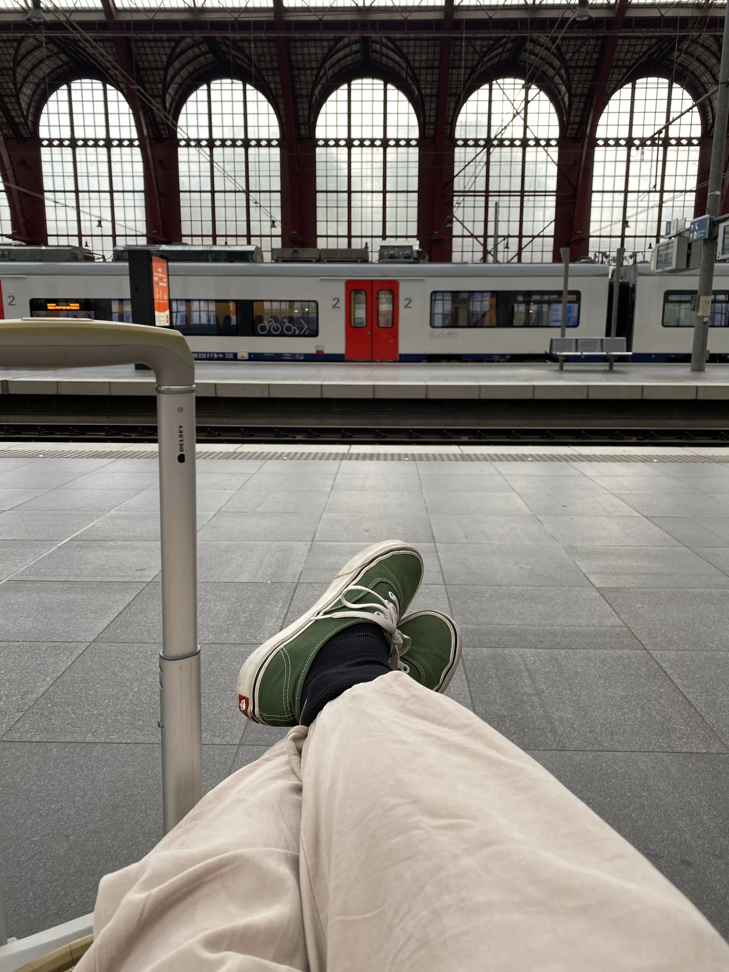 Passenger sitting on a bench at a train station with legs stretched out, wearing beige pants, black socks, and green sneakers. In front, a train with a gray body, red doors, and bicycle symbols on the windows is visible behind the platform.