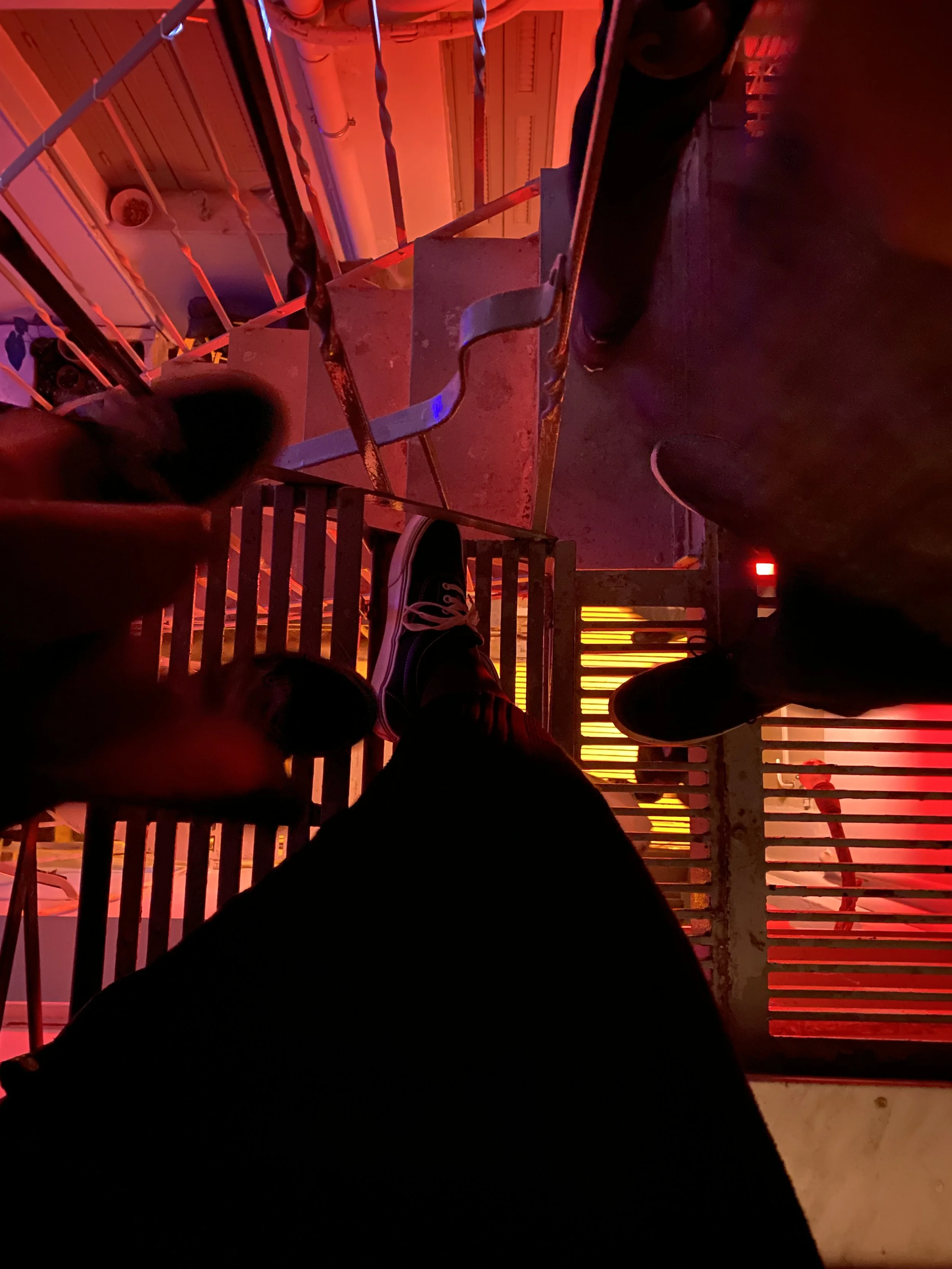 View from a roller coaster at night with red lighting. You see the feet of a person wearing black sneakers, standing on a metal platform, with the coaster track and a red wall in the background.