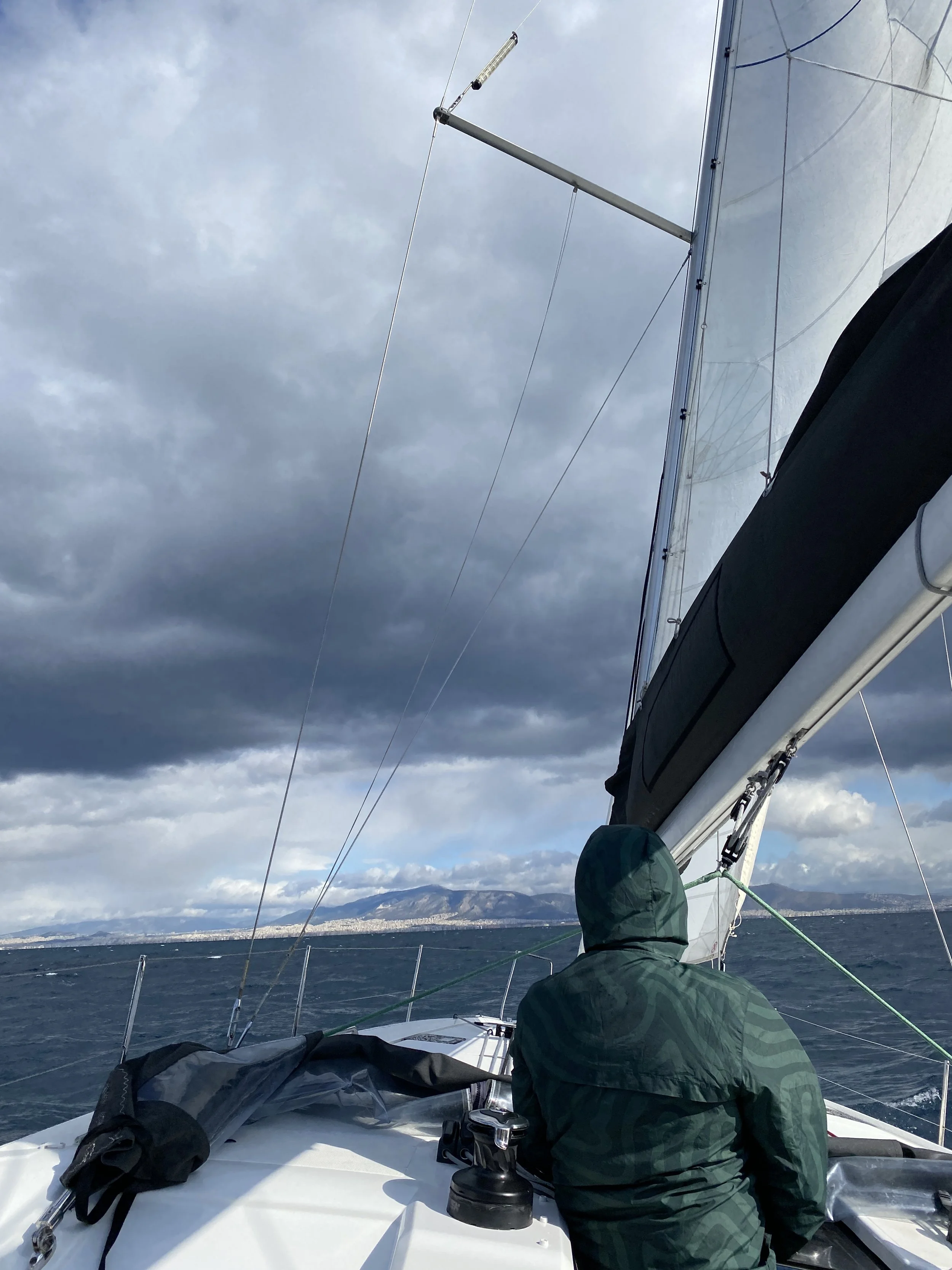 Person in camouflage jacket sitting on sailboat, facing open water with mountains and cloudy sky in the background.