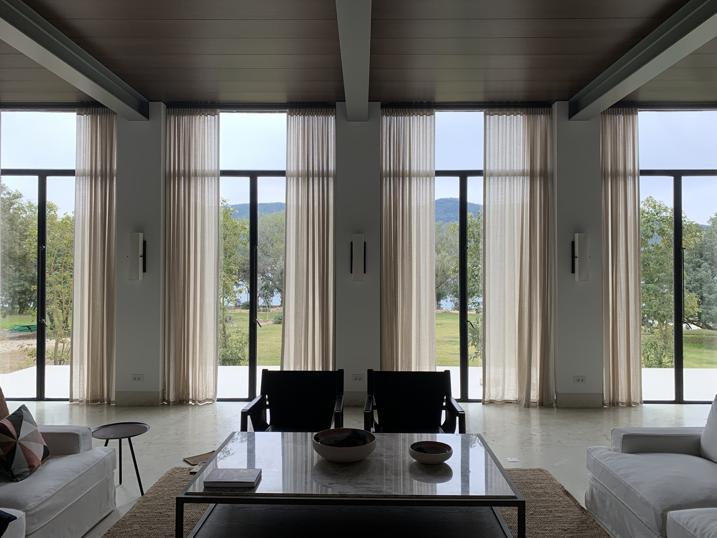 Living room with large floor-to-ceiling windows and beige curtains, overlooking trees and mountains in the distance. There are couches, a coffee table with several bowls, and a chair in the foreground.