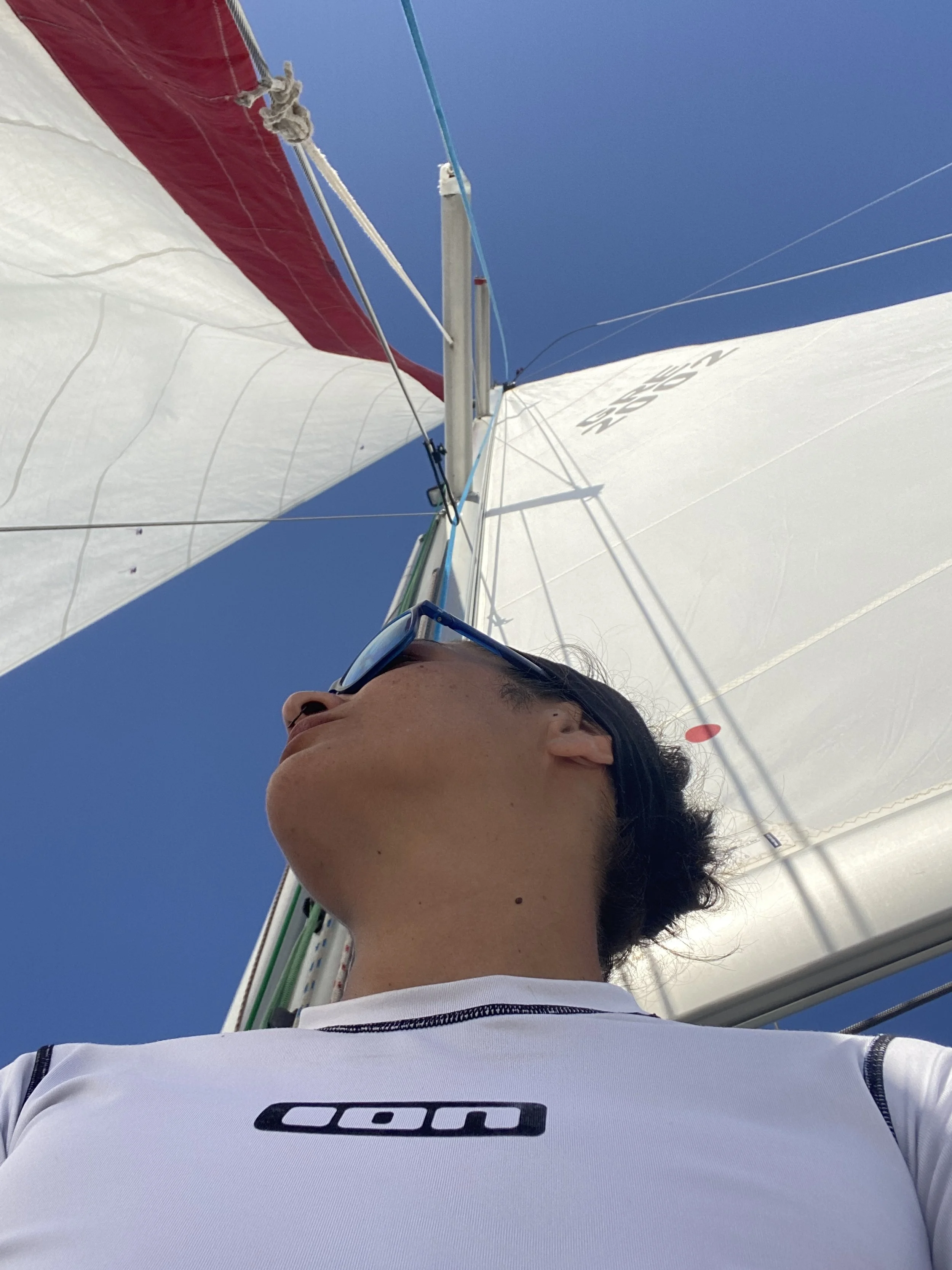 A person wearing sunglasses and a white shirt looks up at the sails of a sailboat against a clear blue sky.