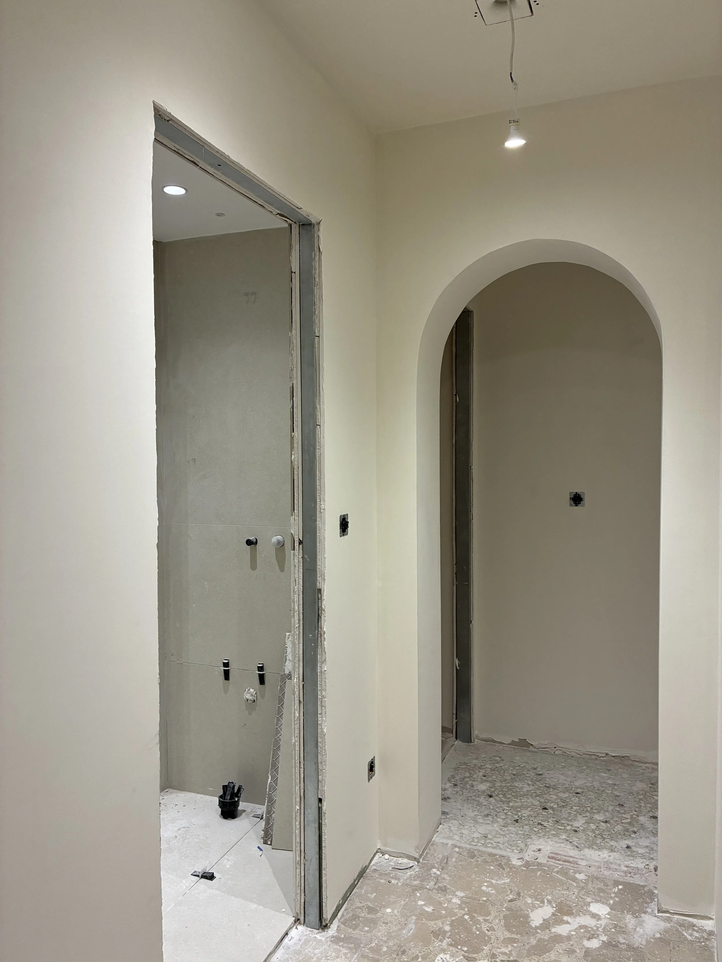 Interior view of a house under construction, showing a doorway to a bathroom with incomplete wall fixtures and a tiled floor, and an arched doorway leading to another room.